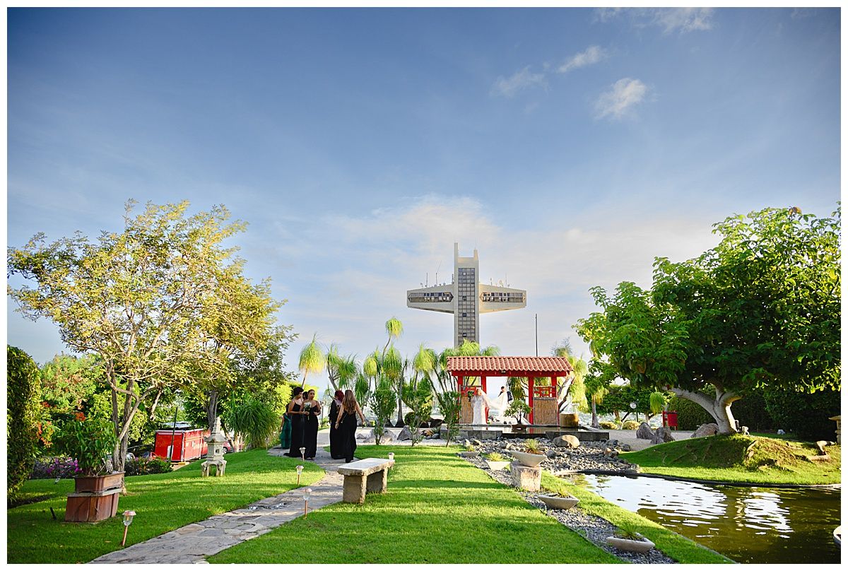 Lush green garden with a large white cross monument, red pavilion, and manicured lawn under a blue sky.