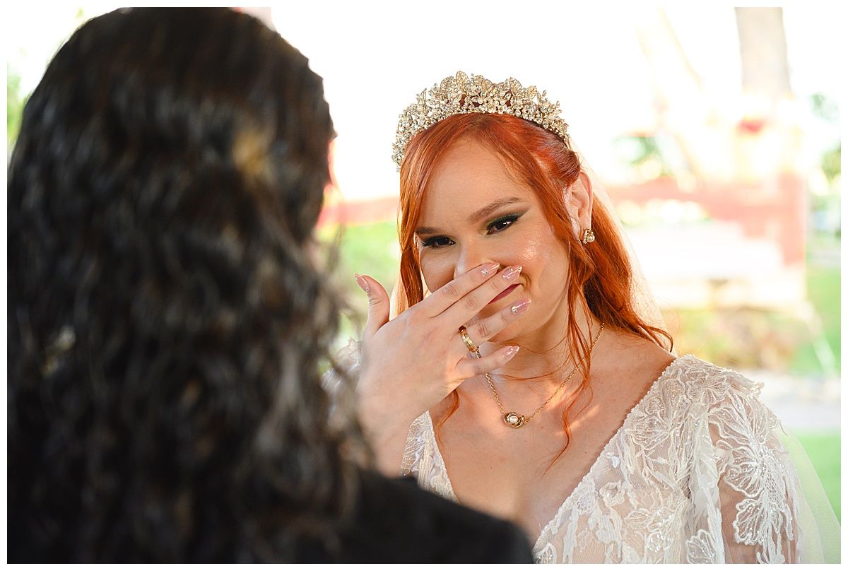 Emotional bride in lace gown and crystal tiara covers her mouth with ringed hand during heartfelt wedding ceremony moment.