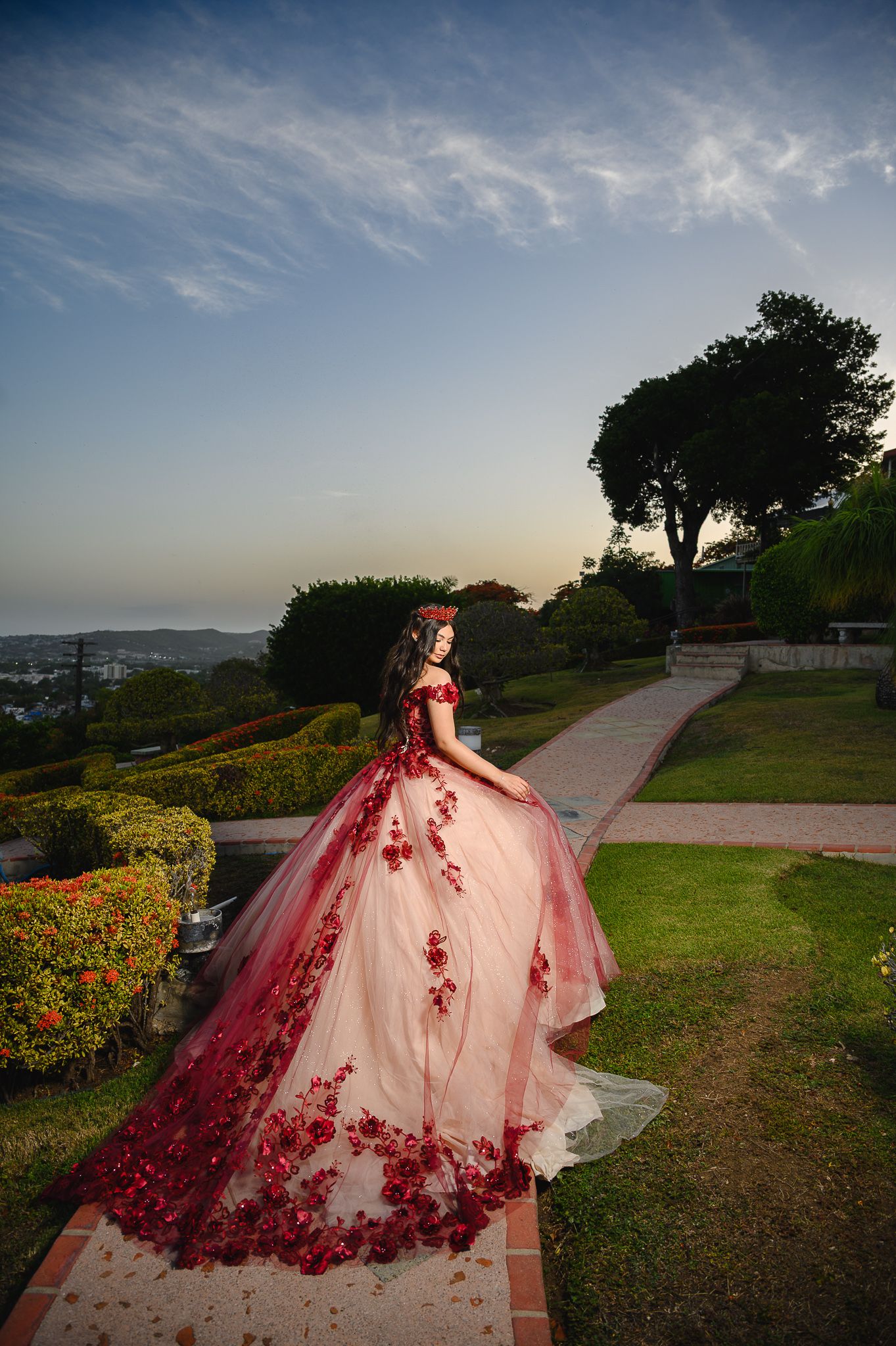 Young woman in stunning red floral ball gown poses gracefully in lush garden at sunset.