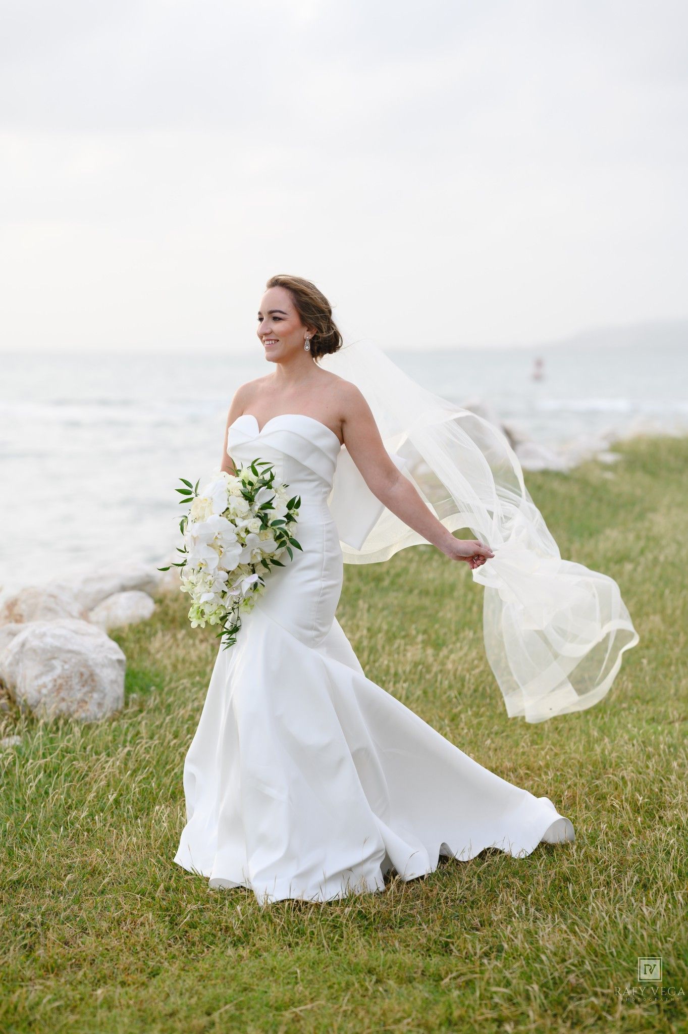 Bride in strapless white mermaid gown holds bouquet, veil flowing in wind by rocky lakeside.