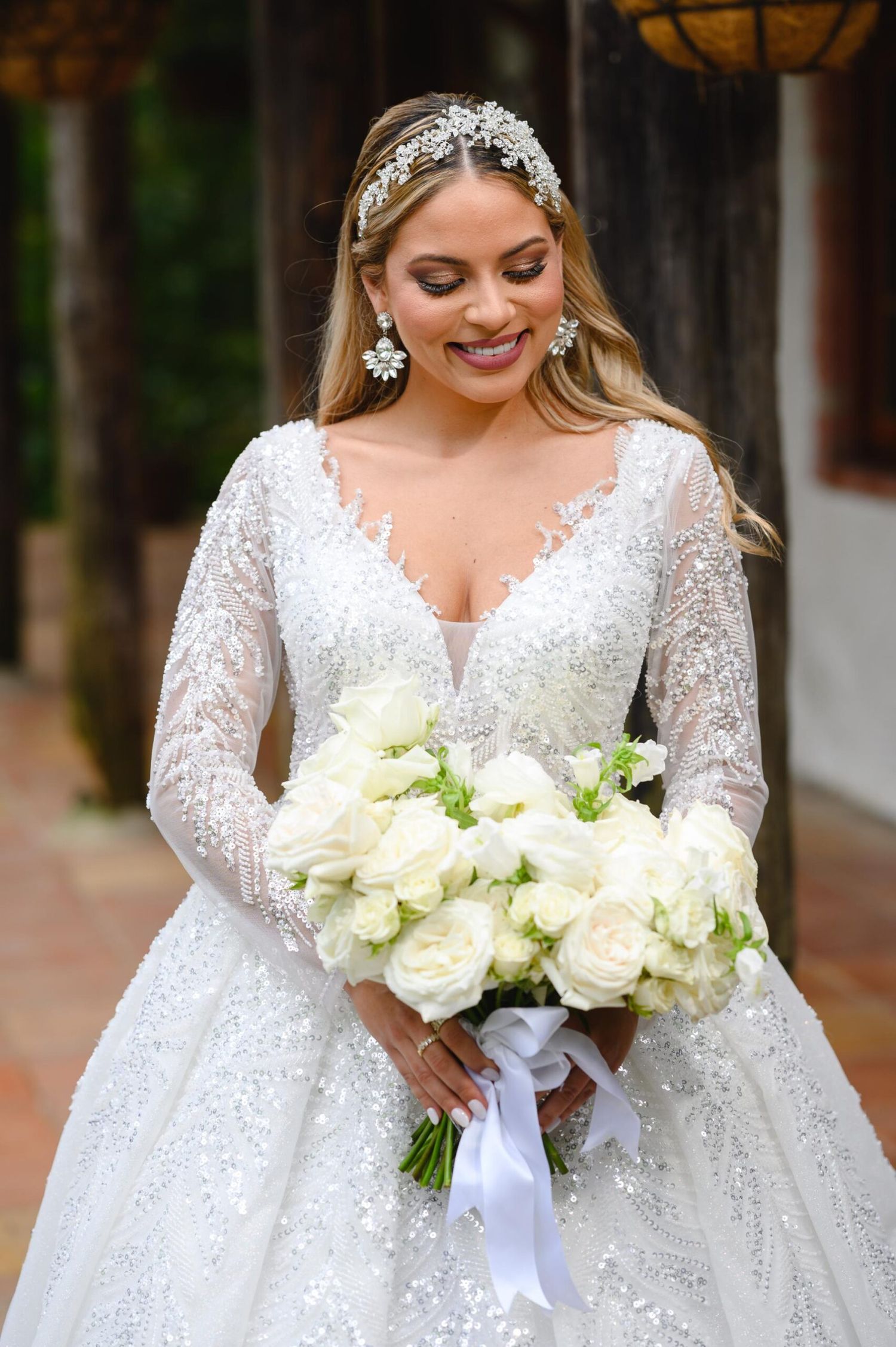 Bride in beaded long-sleeve wedding gown holding white rose bouquet, wearing crystal headpiece and earrings, smiling.