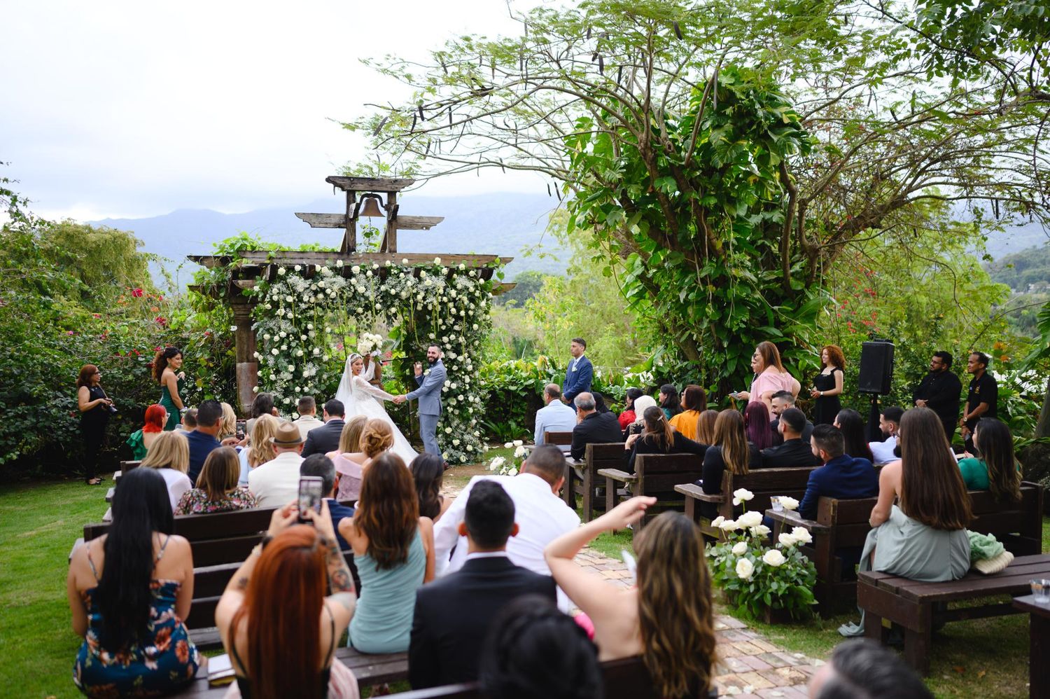 Outdoor wedding ceremony with guests seated on wooden benches, floral arch, lush greenery, and scenic mountain views.