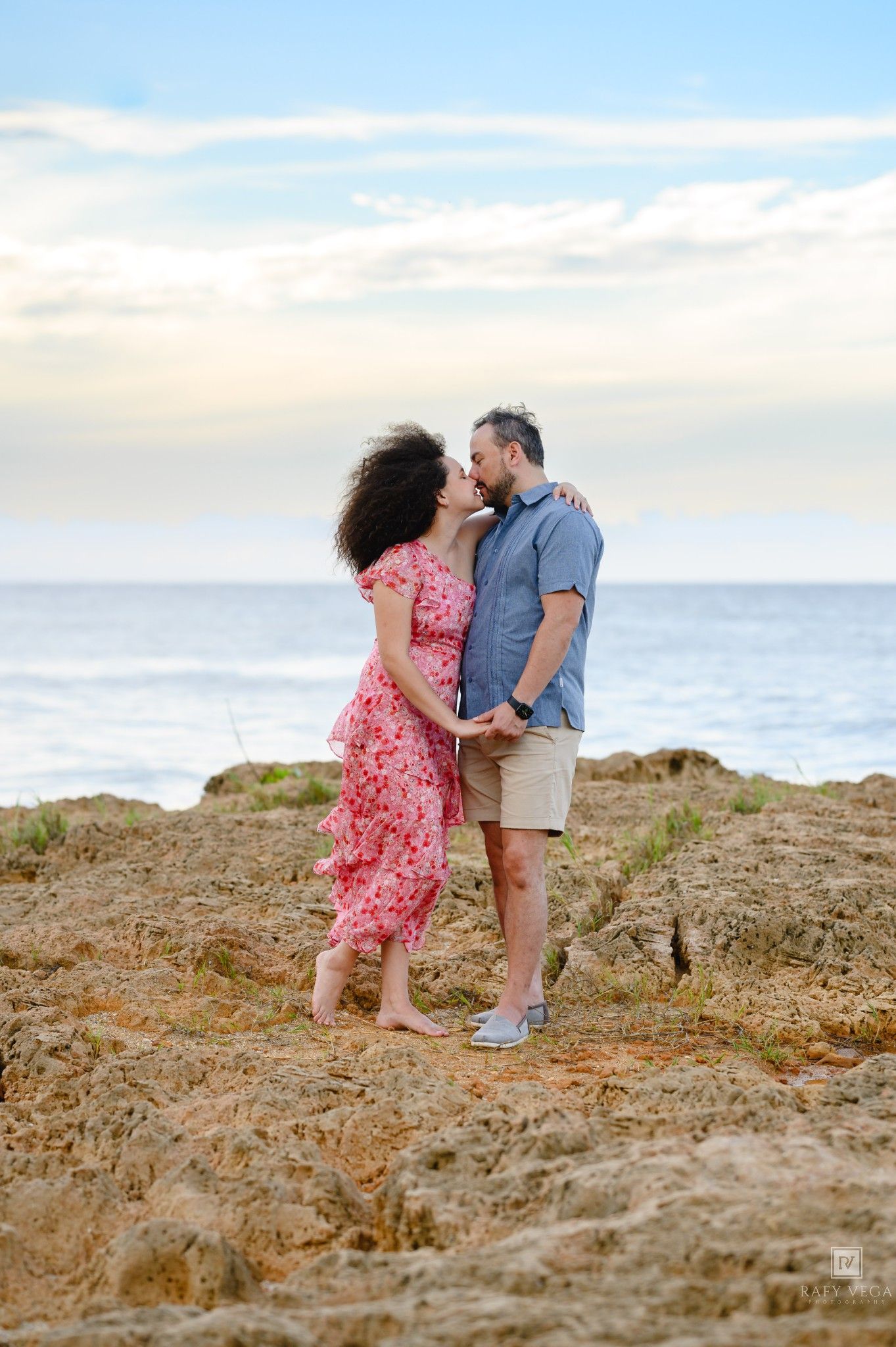 Couple sharing a romantic kiss on rocky coastal cliffs with ocean and pastel sky backdrop.