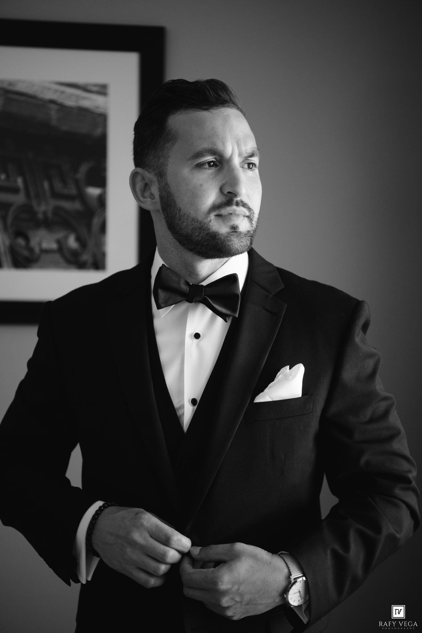 Groom in black tuxedo with bow tie and pocket square buttoning jacket in black and white portrait photo.