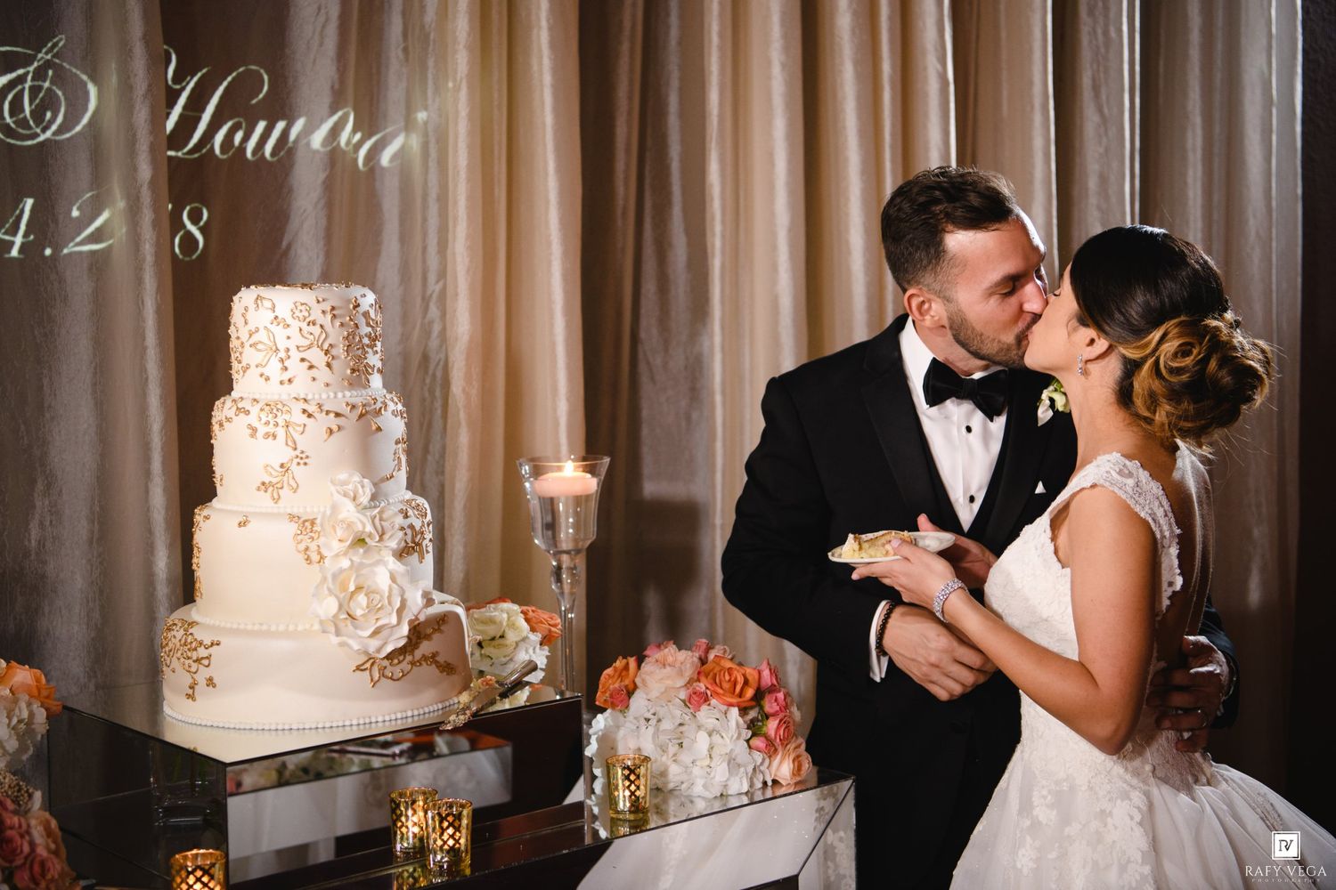 Bride and groom sharing a kiss beside a elegant white and gold tiered wedding cake at their reception.