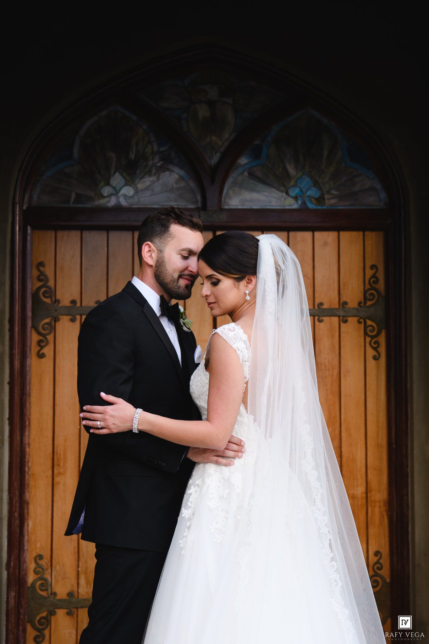 Bride and groom share romantic moment in front of ornate wooden church doors with stained glass window.