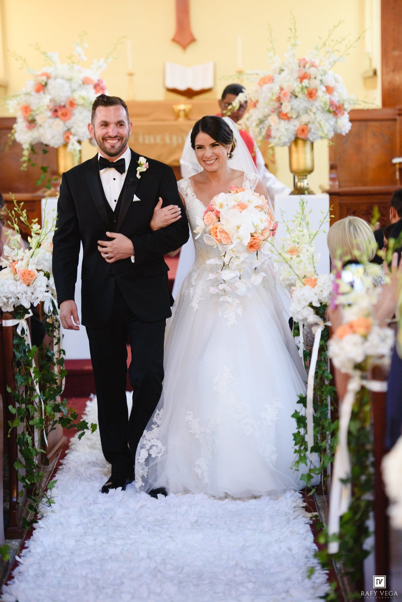 Bride and groom walking down church aisle after wedding ceremony, surrounded by elegant floral arrangements.