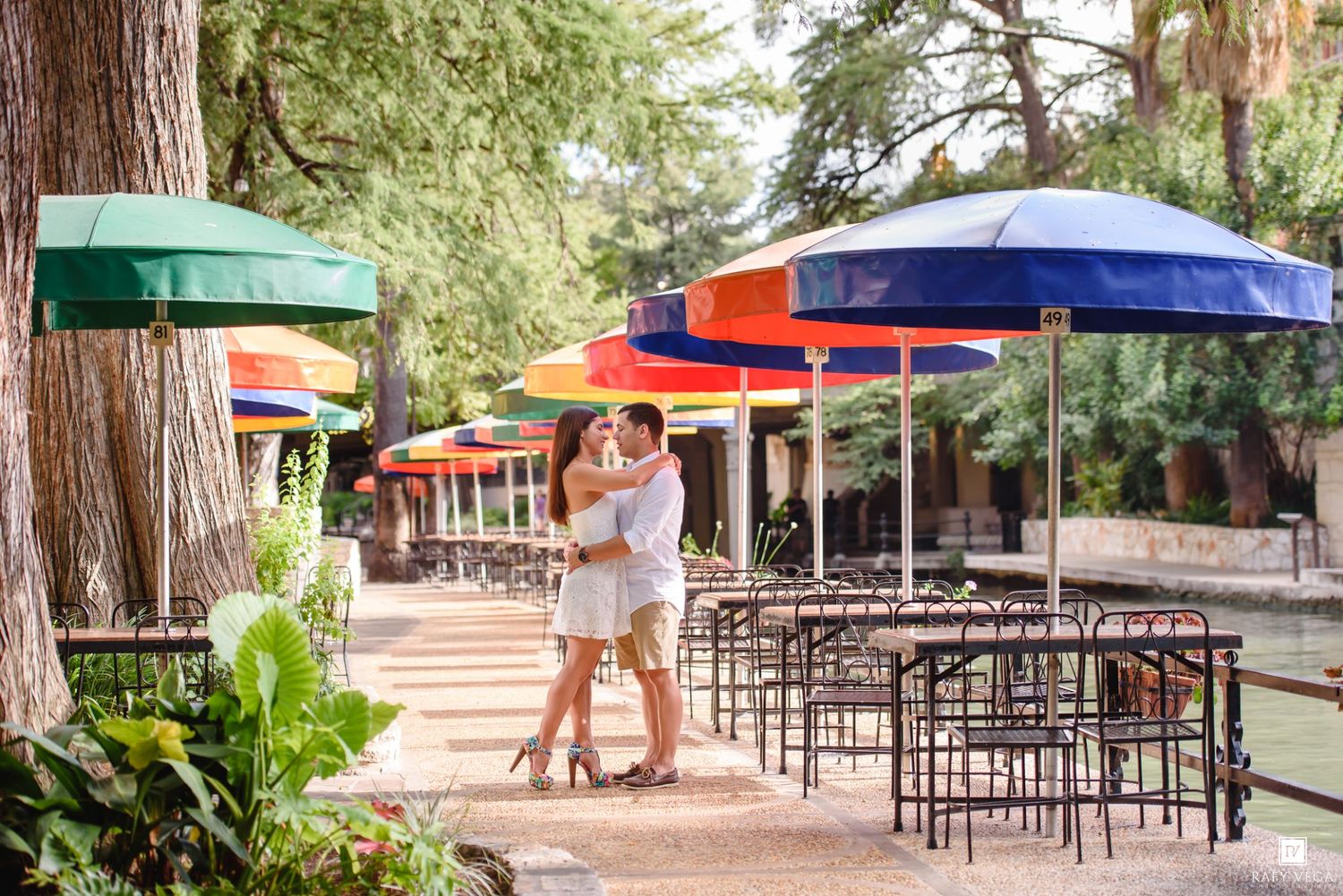 Couple embracing romantically on San Antonio River Walk patio with colorful umbrellas and lush trees.
