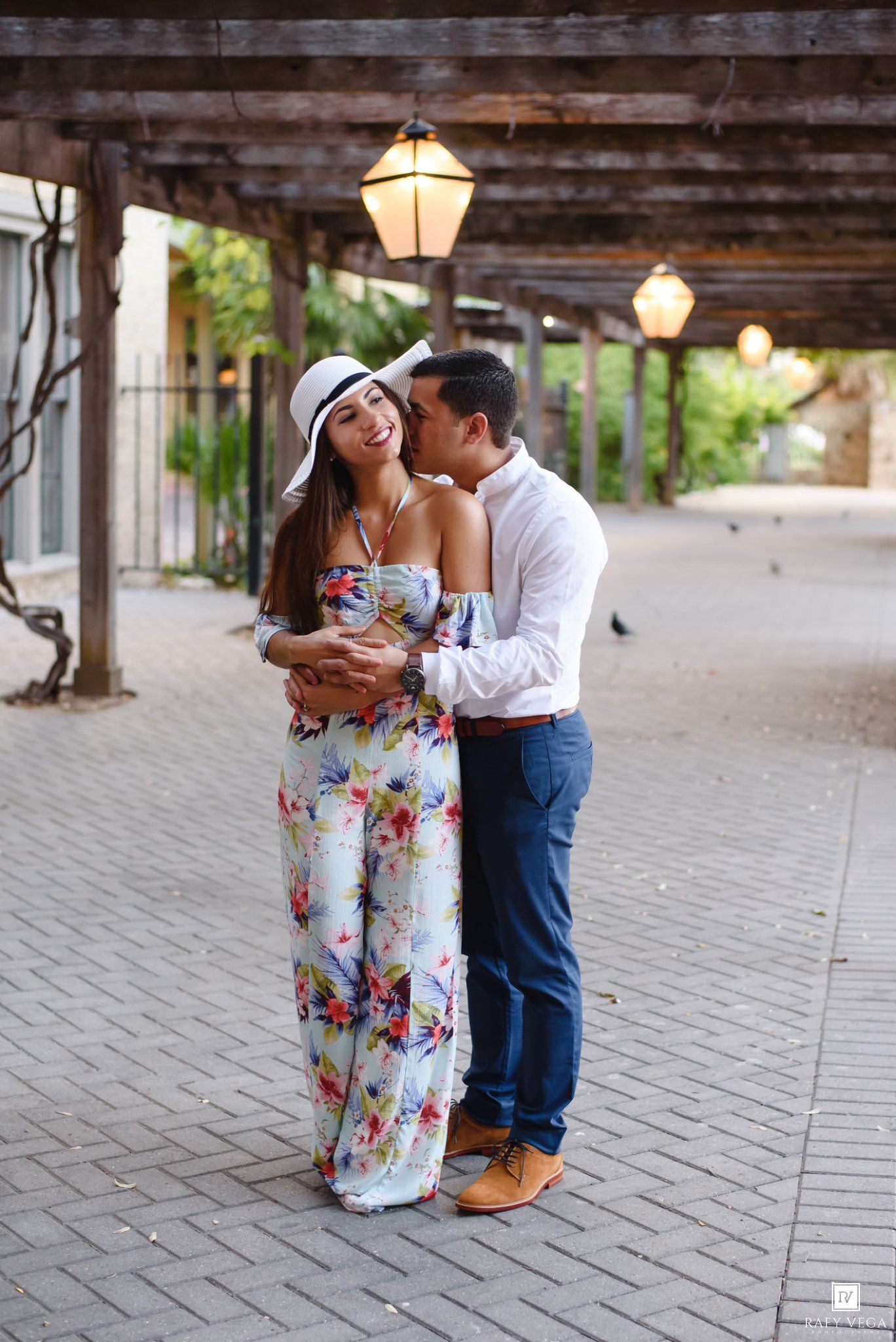 Couple embracing under wooden pergola with lanterns, woman in floral maxi dress and white hat, man in white shirt.