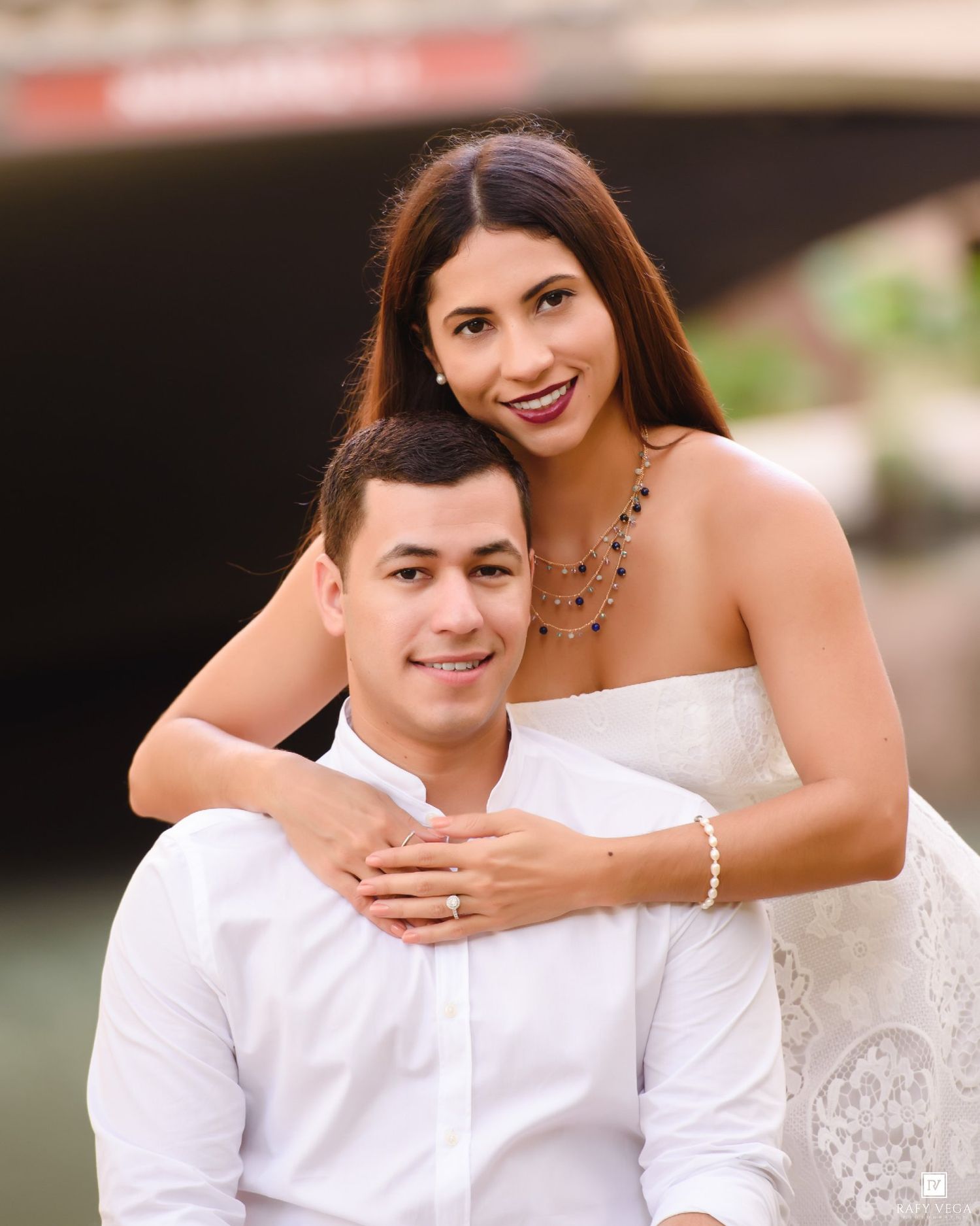Couple posing for engagement photos outdoors near a bridge, woman in white dress with necklace, man in white shirt.