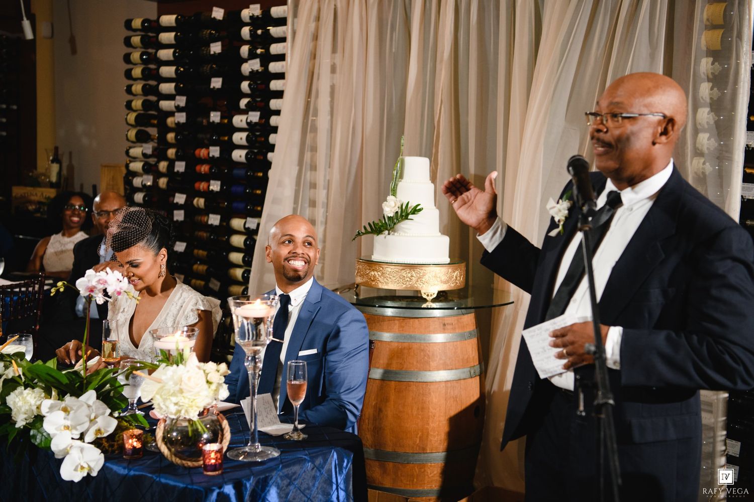 Man giving speech at wedding reception near wine barrel cake stand, couple seated at decorated table.
