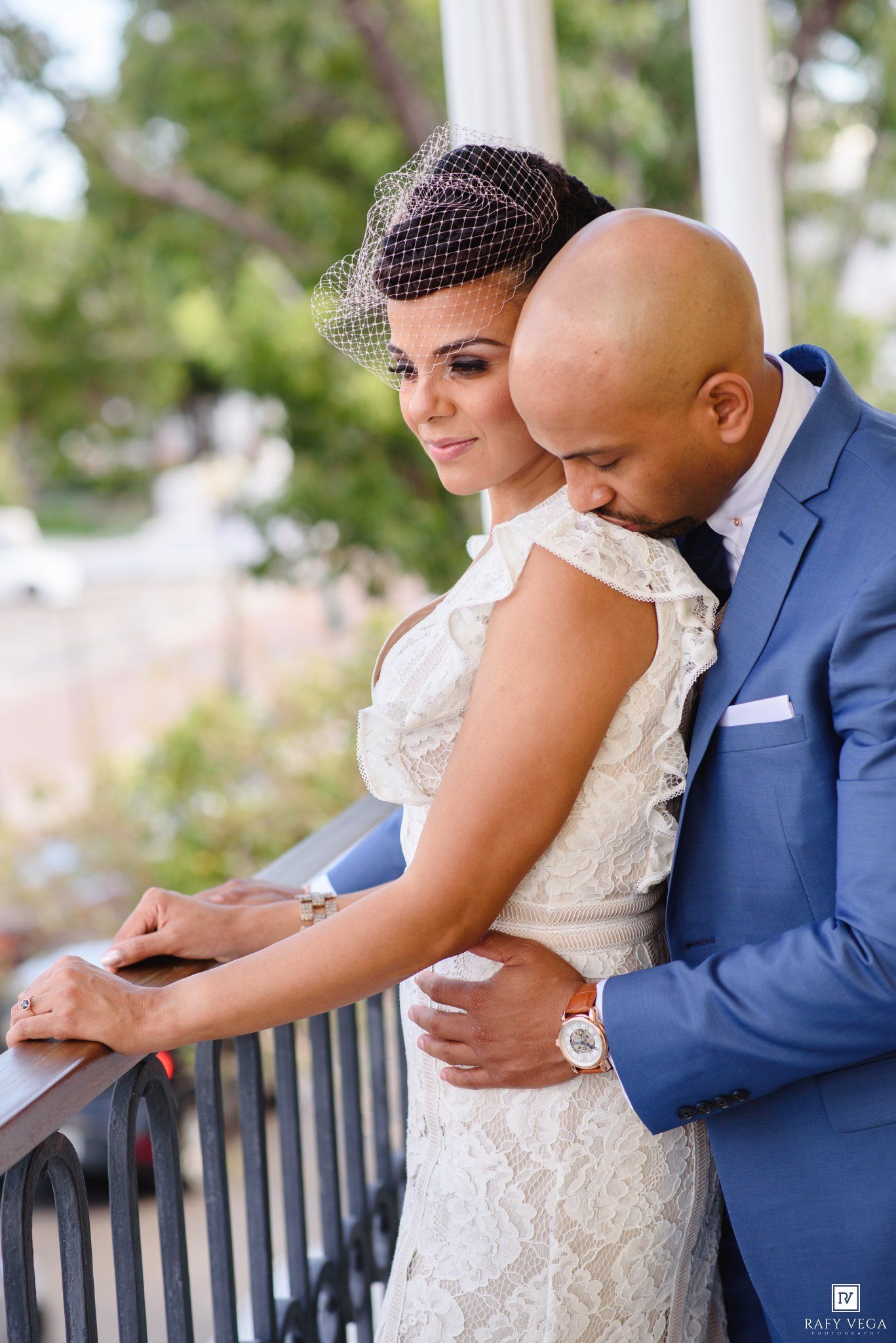 Bride and groom share a romantic embrace on a balcony, bride in lace dress with birdcage veil, groom in blue suit.