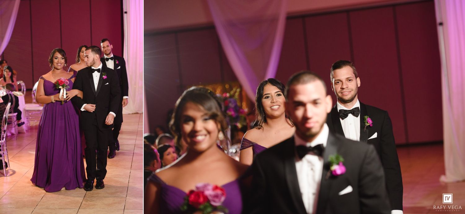 Wedding party members in formal attire walk into a reception hall with pink draping and decorations.