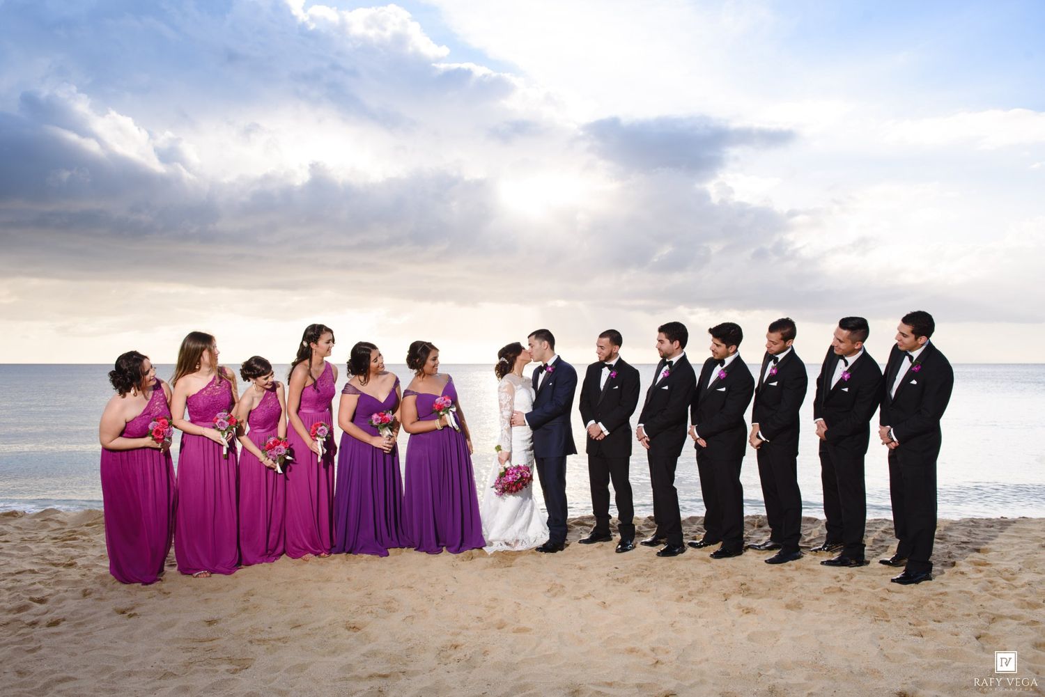 Wedding party on beach, bridesmaids in purple gowns and groomsmen in black suits flanking bride and groom.