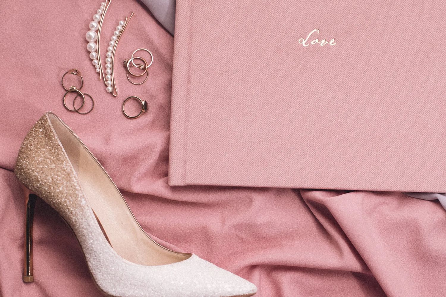 Bridal flat lay with glitter heel, pearl necklace, gold rings, and pink velvet love journal.