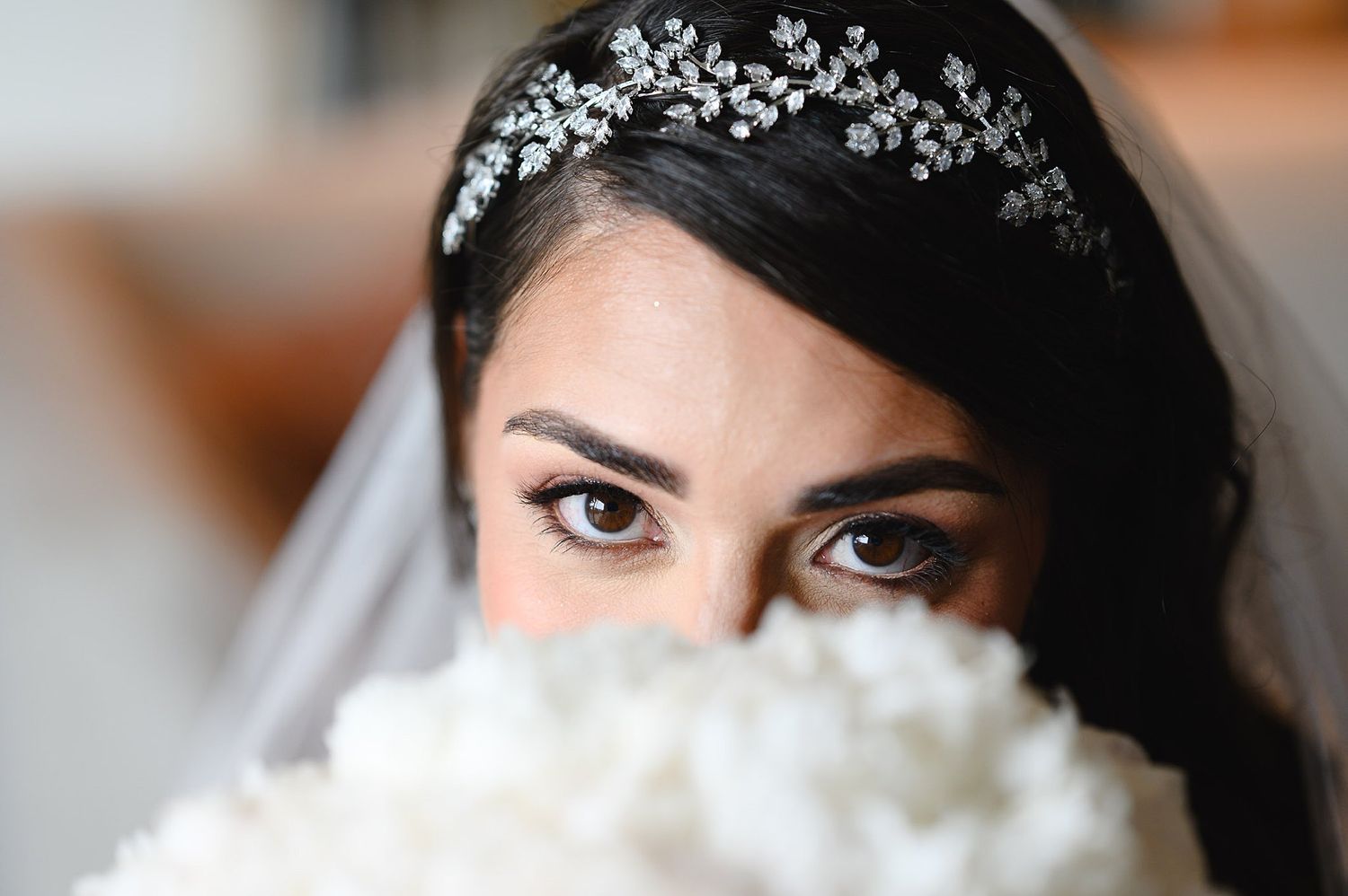 Bride with dark hair wearing a floral crystal tiara and veil, holding a white bouquet up to her face.
