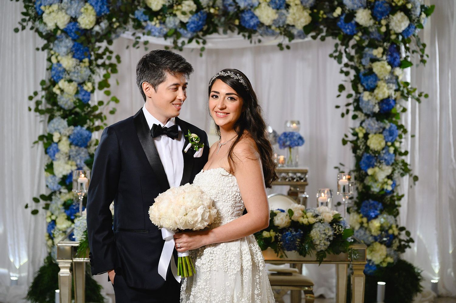 Bride and groom pose under a blue and white floral arch at their elegant wedding ceremony.