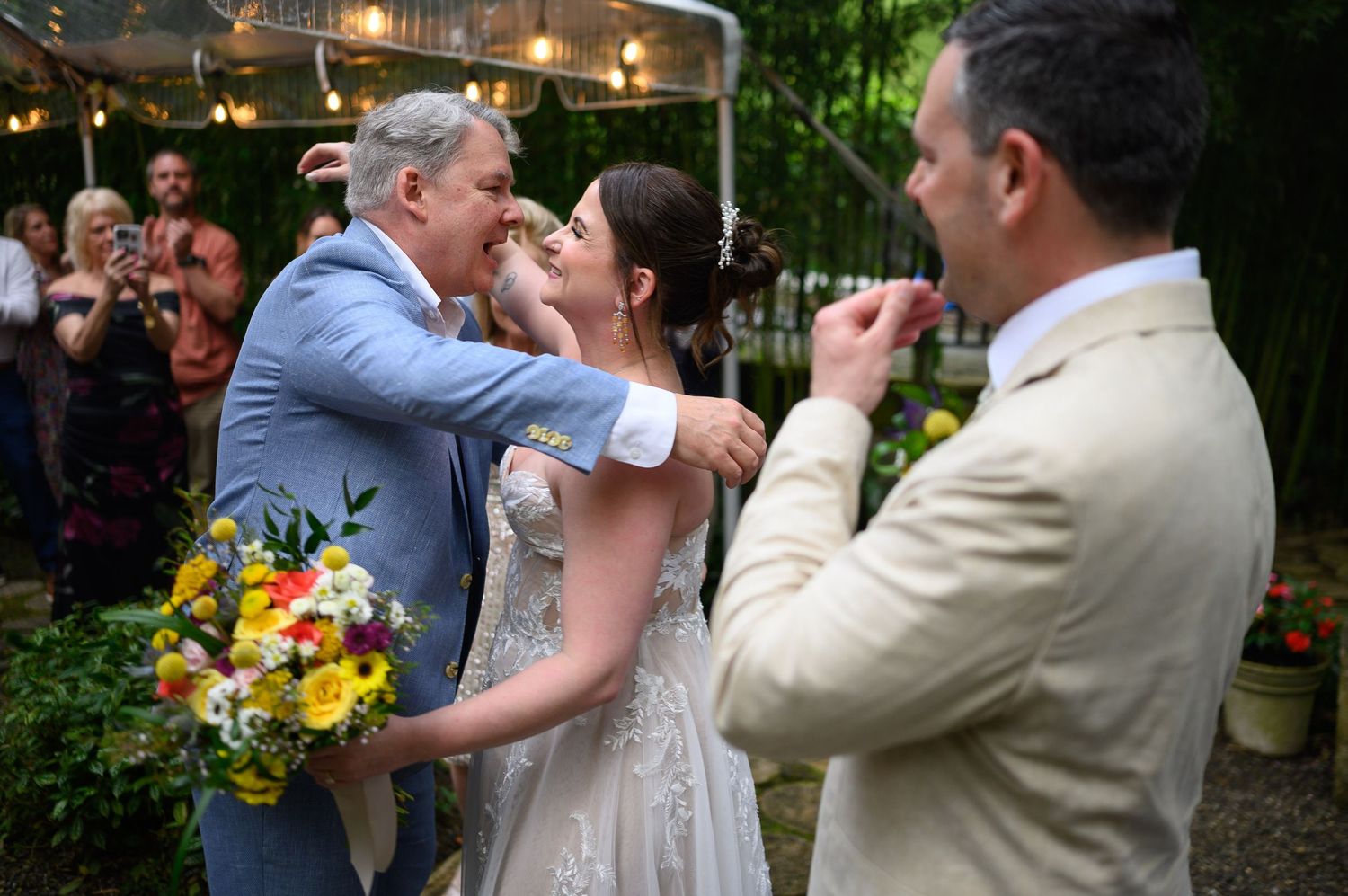 Bride in white gown receives congratulations from older man in blue suit at outdoor wedding ceremony.