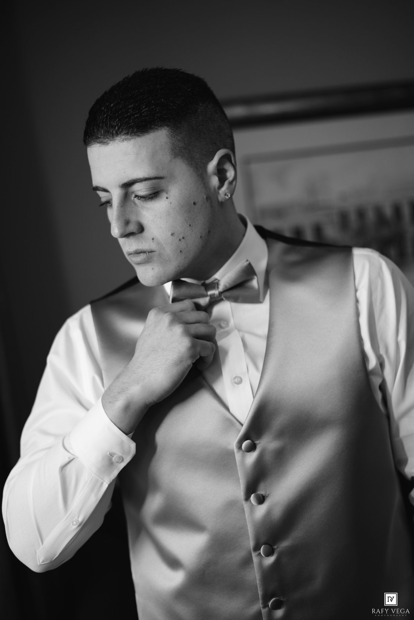 Young groom adjusting bow tie wearing white dress shirt and vest in black and white wedding photo.