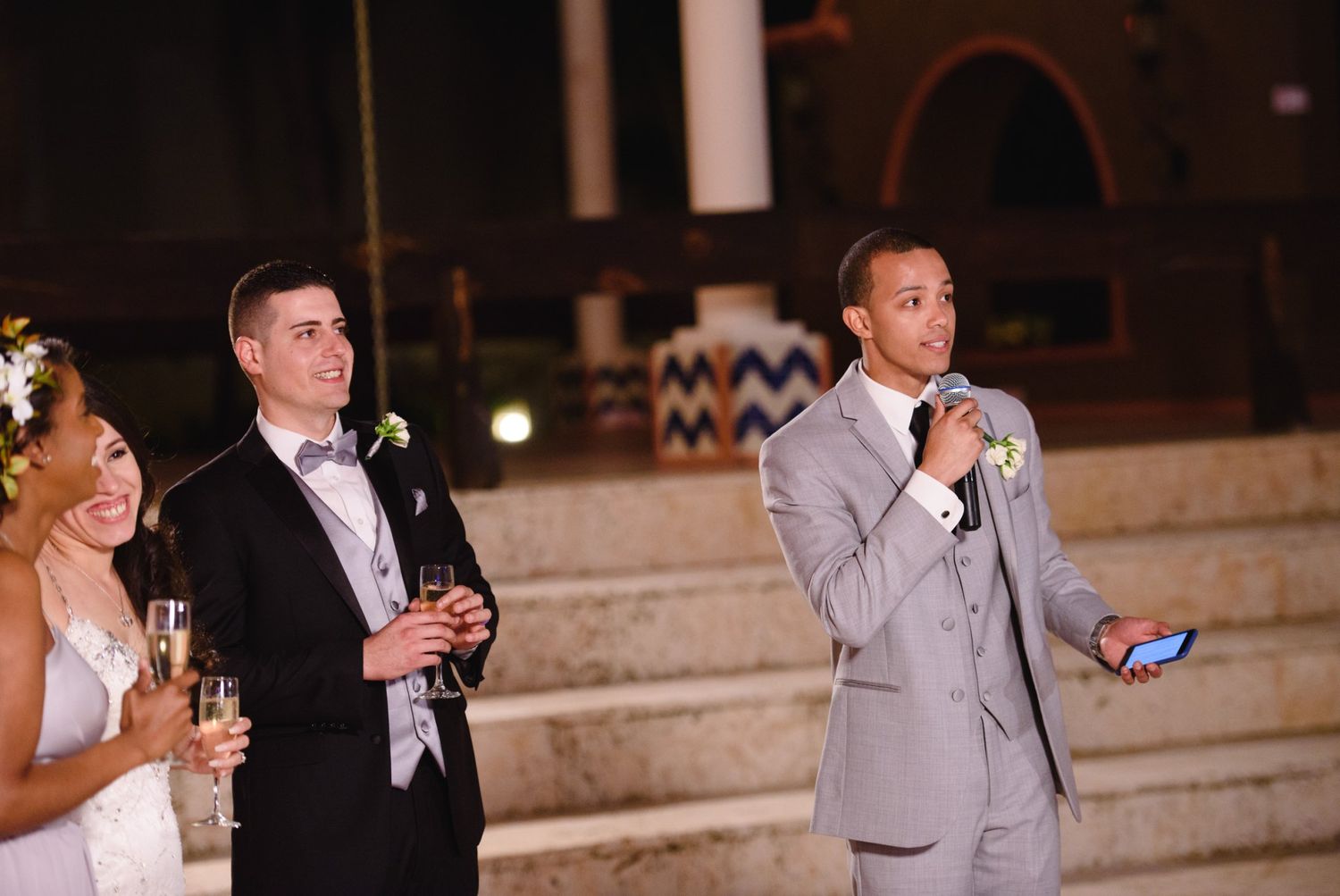 Groom in gray suit gives wedding speech with microphone while bride and groom in tuxedo smile holding champagne.