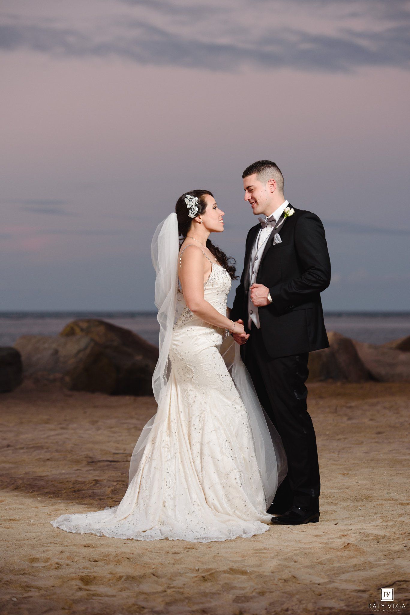 Bride and groom holding hands on sandy beach at sunset, bride in lace mermaid gown with veil, groom in black tuxedo.
