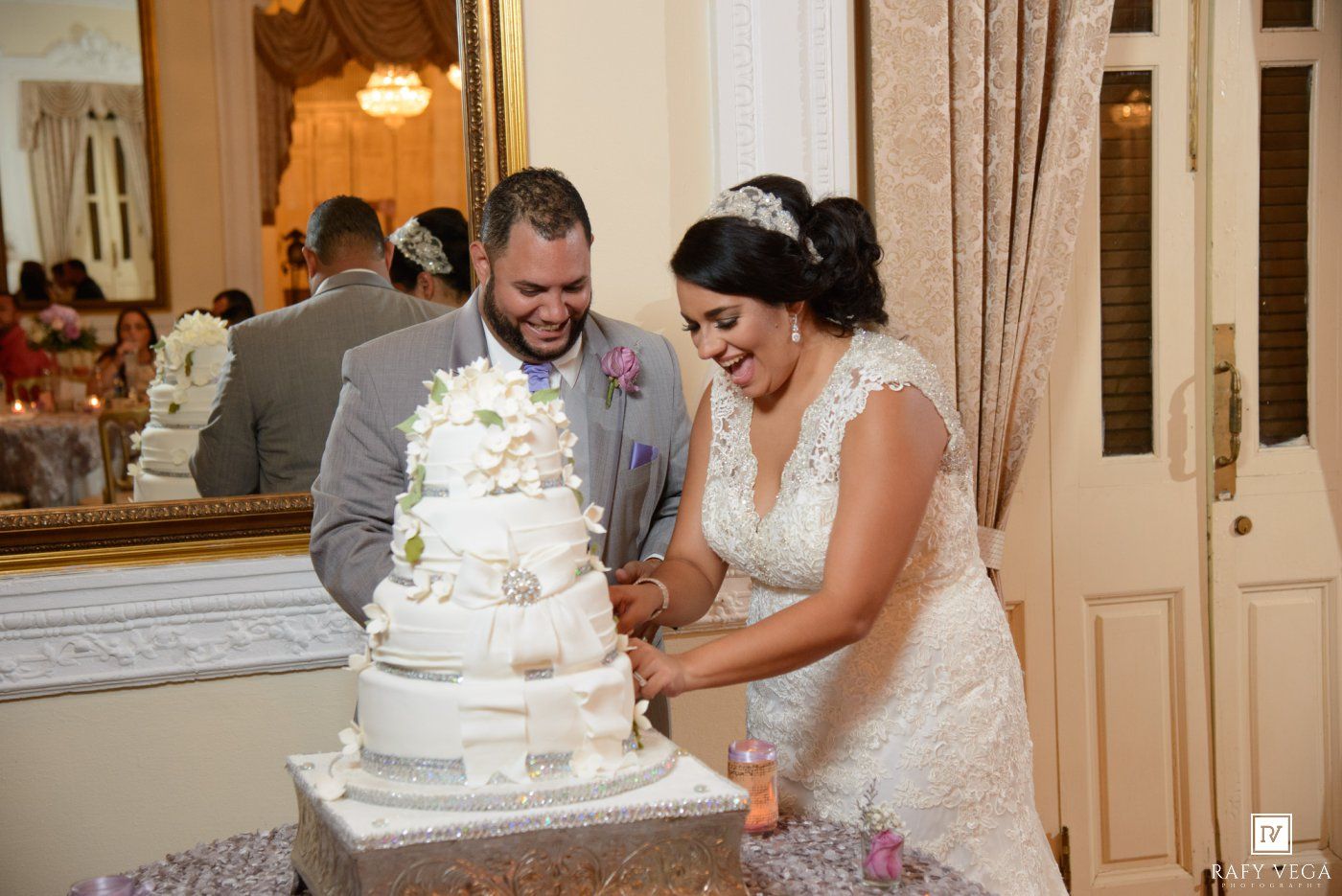Bride and groom smiling while cutting their white tiered wedding cake at elegant reception venue.
