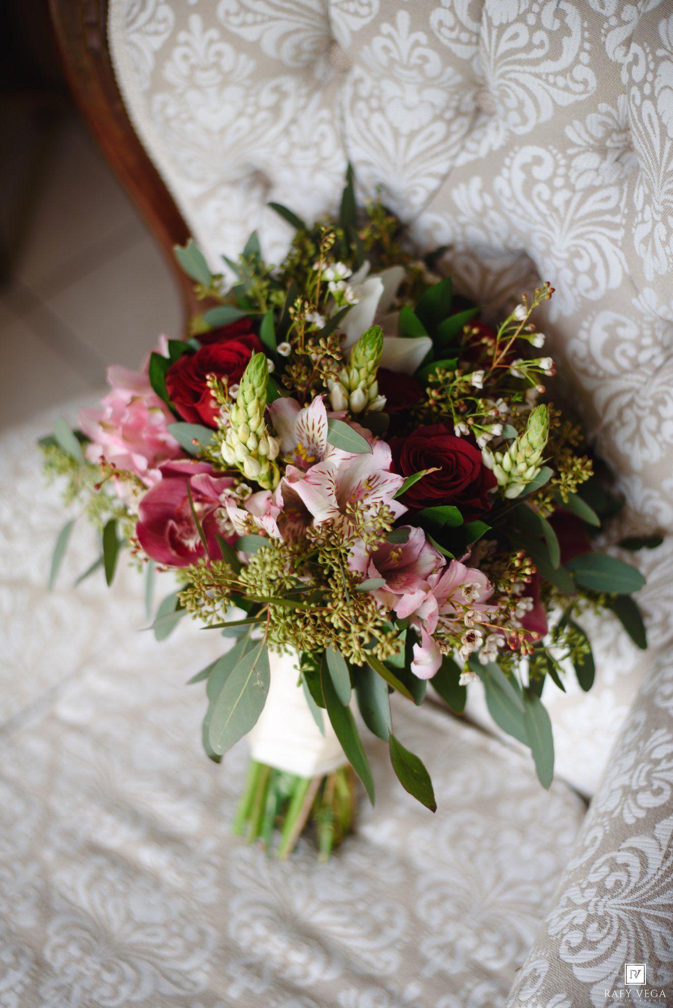 Elegant bridal bouquet with deep red roses, pink alstroemeria, and greenery resting on ornate white chair.
