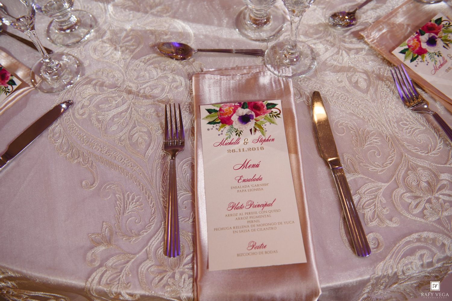 Close-up of a wedding place setting with floral menu card, silverware, and crystal glasses on embossed white linen.