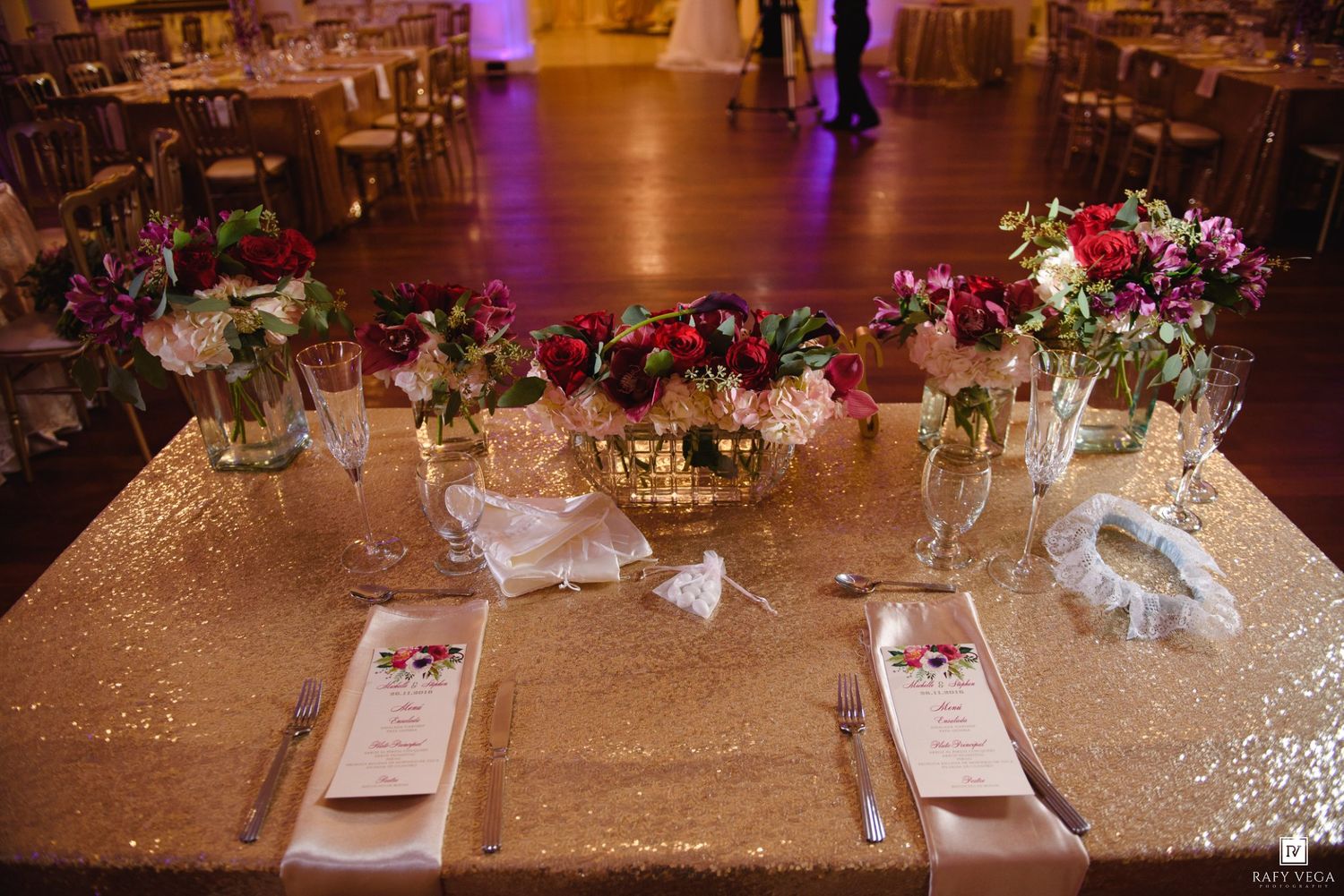 Elegant wedding reception head table with gold sequin tablecloth, floral centerpieces, and neatly arranged place settings.