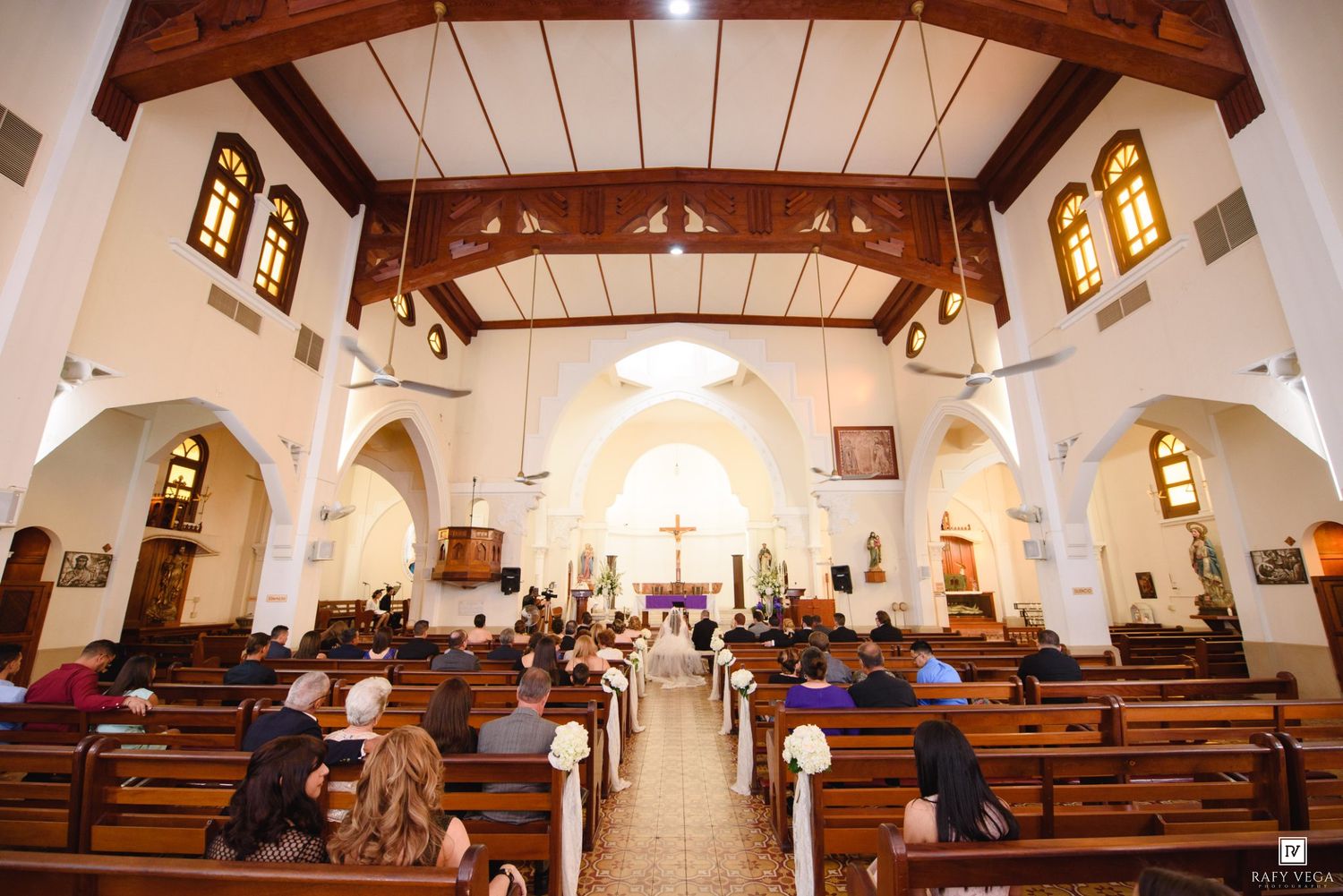 Guests seated in wooden pews inside a white-walled Catholic church with wooden ceiling during a ceremony.