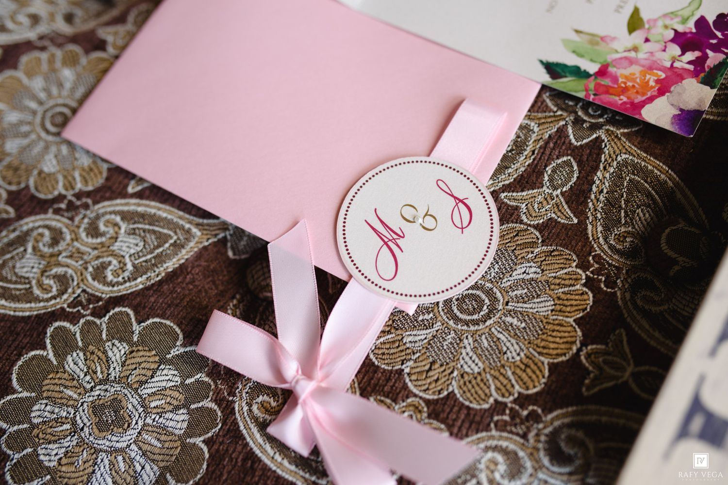 Pink wedding invitation with monogram tag and satin ribbon bow on floral patterned fabric background.