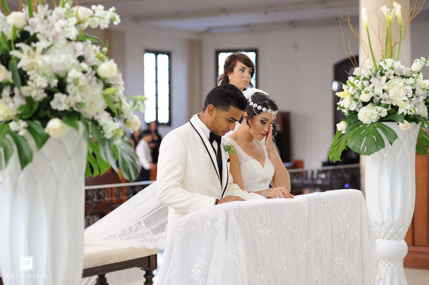Bride and groom bow heads in prayer at elegant church wedding altar adorned with white floral arrangements.