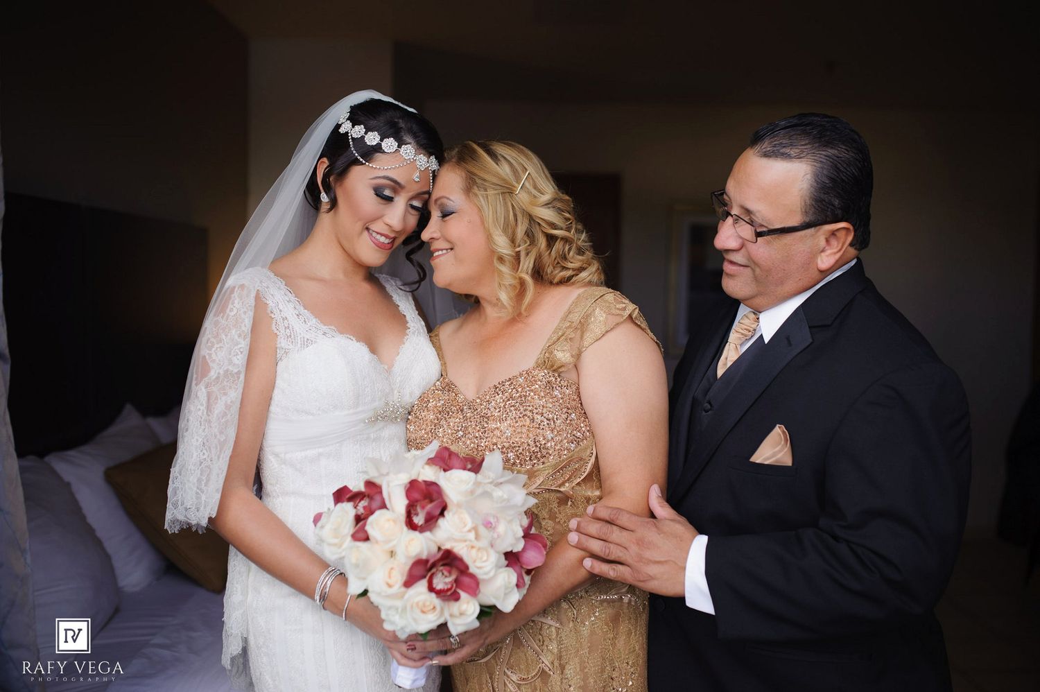 Bride in white lace gown holding bouquet smiles with parents in elegant attire during wedding day preparations.