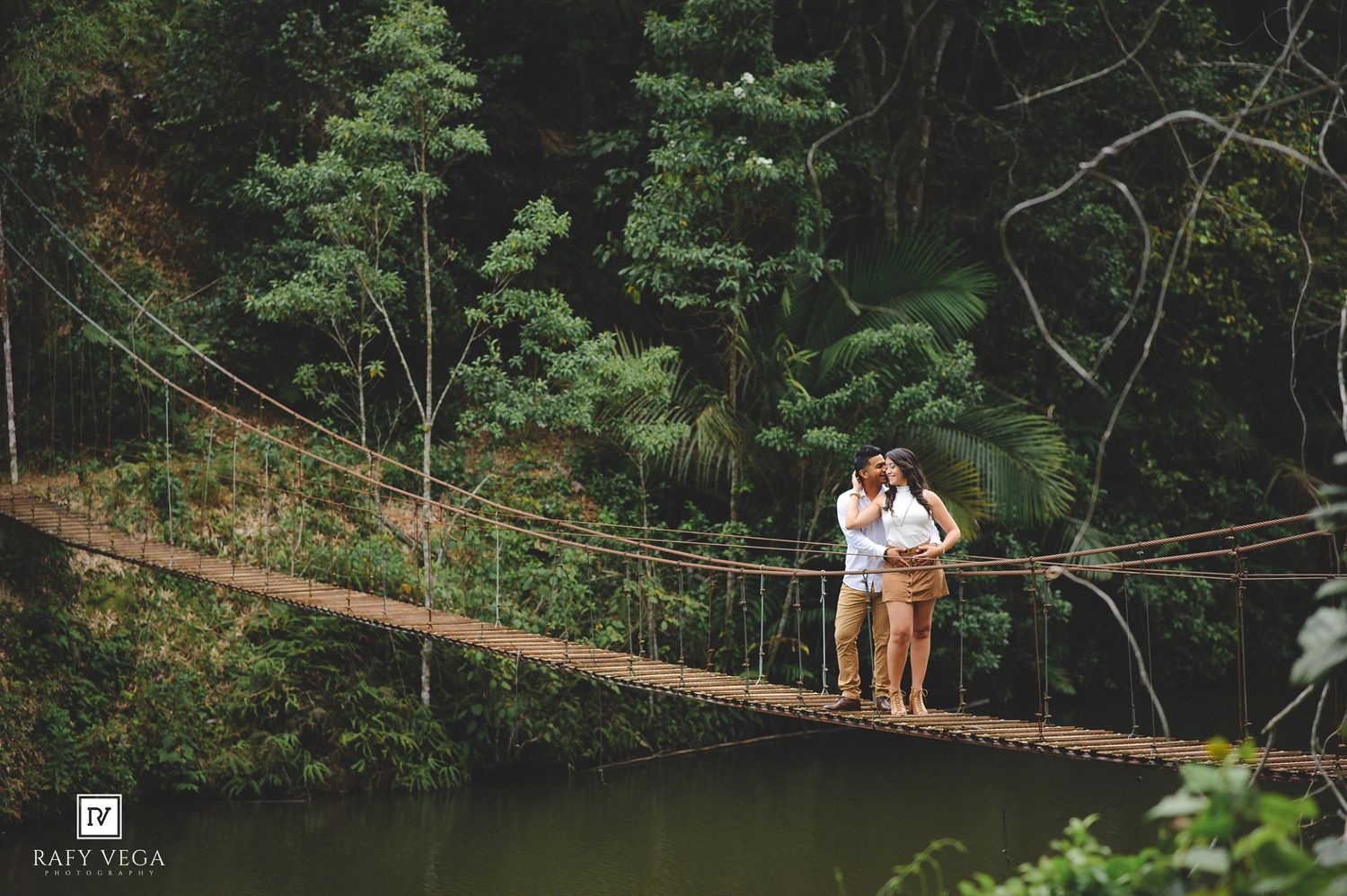 Couple embracing on a rustic rope suspension bridge surrounded by lush tropical rainforest trees.