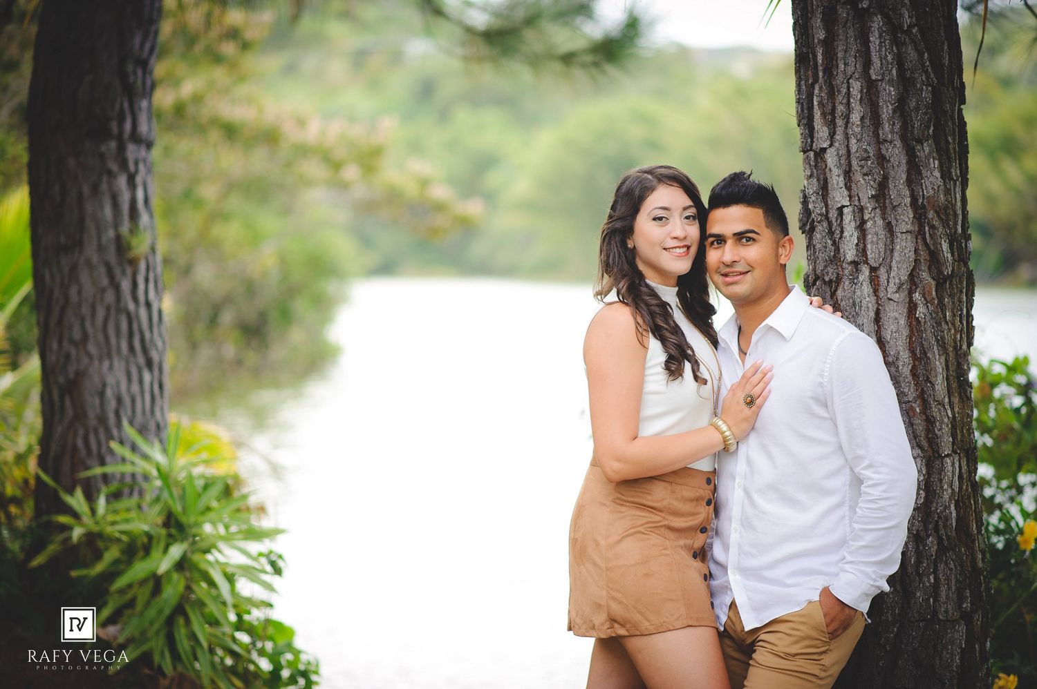 Couple posing romantically by a lake surrounded by trees, woman in white top and tan skirt, man in white shirt.