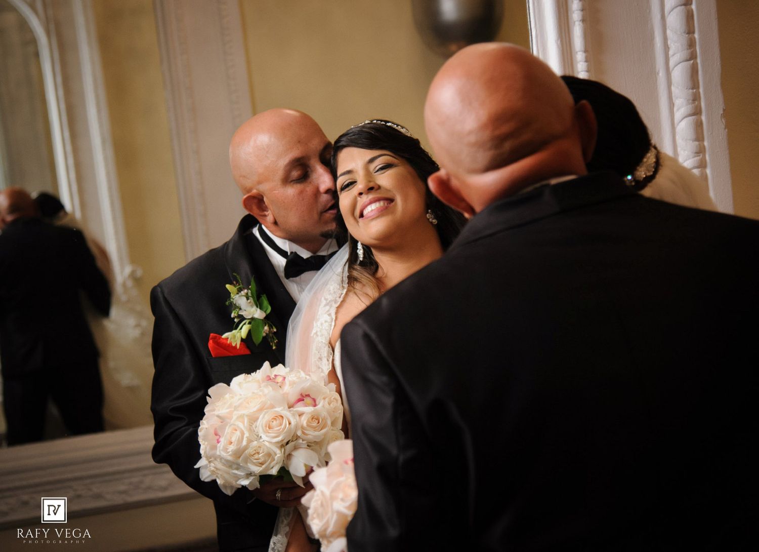 Bride smiles joyfully holding white roses bouquet while groom kisses her cheek during wedding ceremony indoors.
