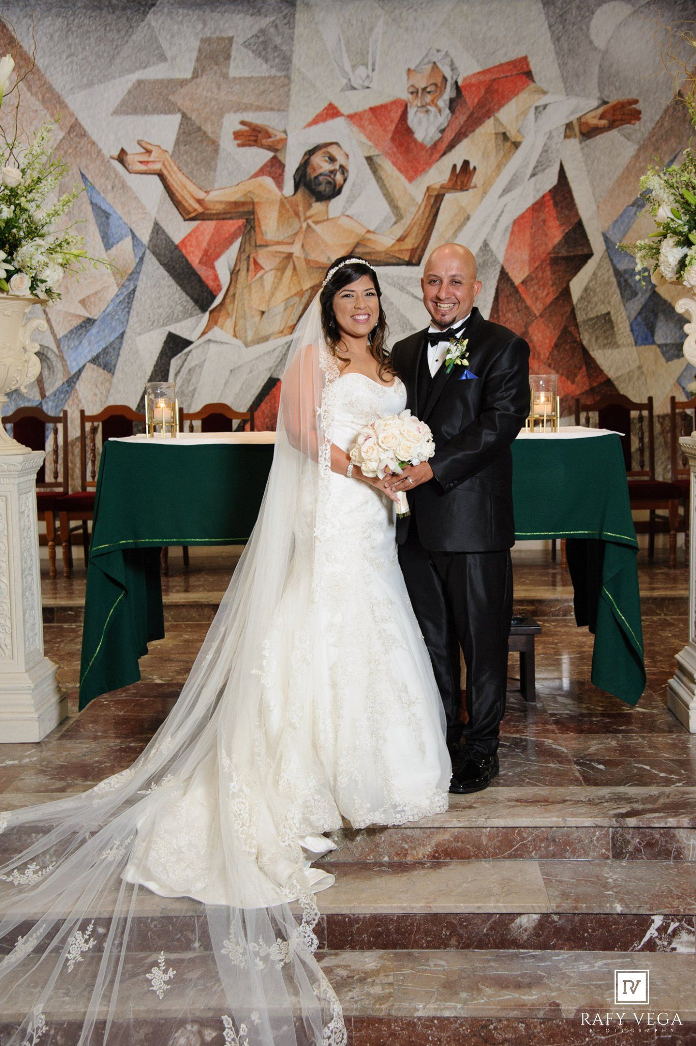 Bride and groom pose together in church before colorful mosaic mural on their wedding day.