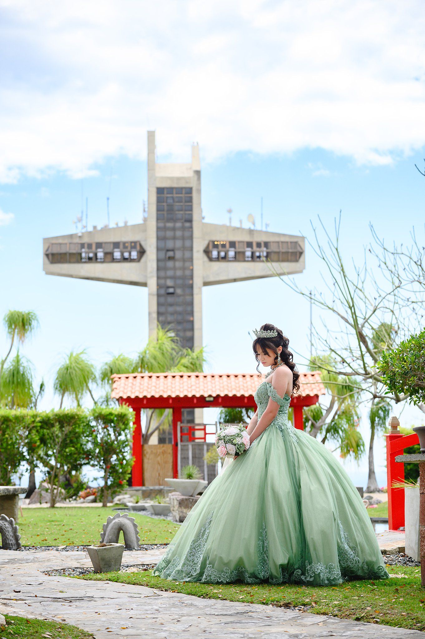 Young woman in elegant green ball gown poses in garden with large cross monument behind her on a sunny day.