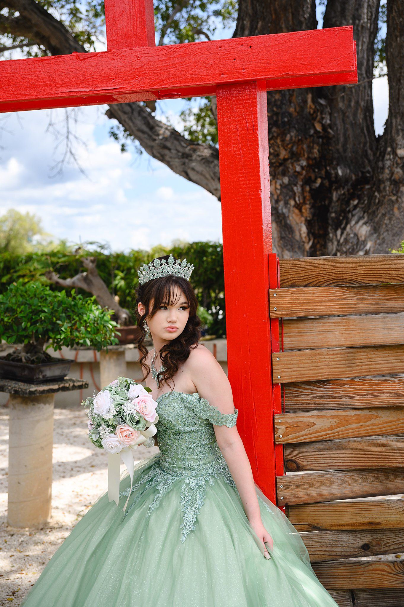 Young woman in sage green quinceañera gown with tiara holds pink bouquet beside red torii gate outdoors.