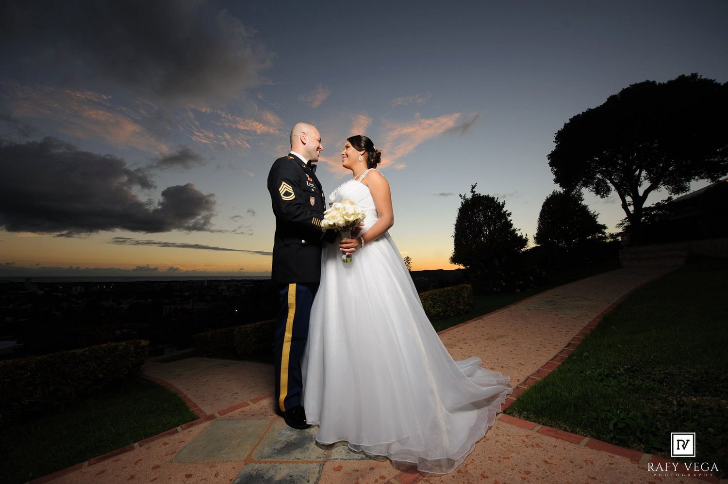 Military groom and bride in white ballgown share romantic moment on garden path at golden sunset.
