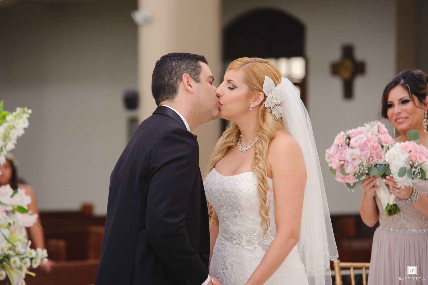 Bride and groom share first kiss at church wedding altar, bridesmaid holding pink bouquet in background.