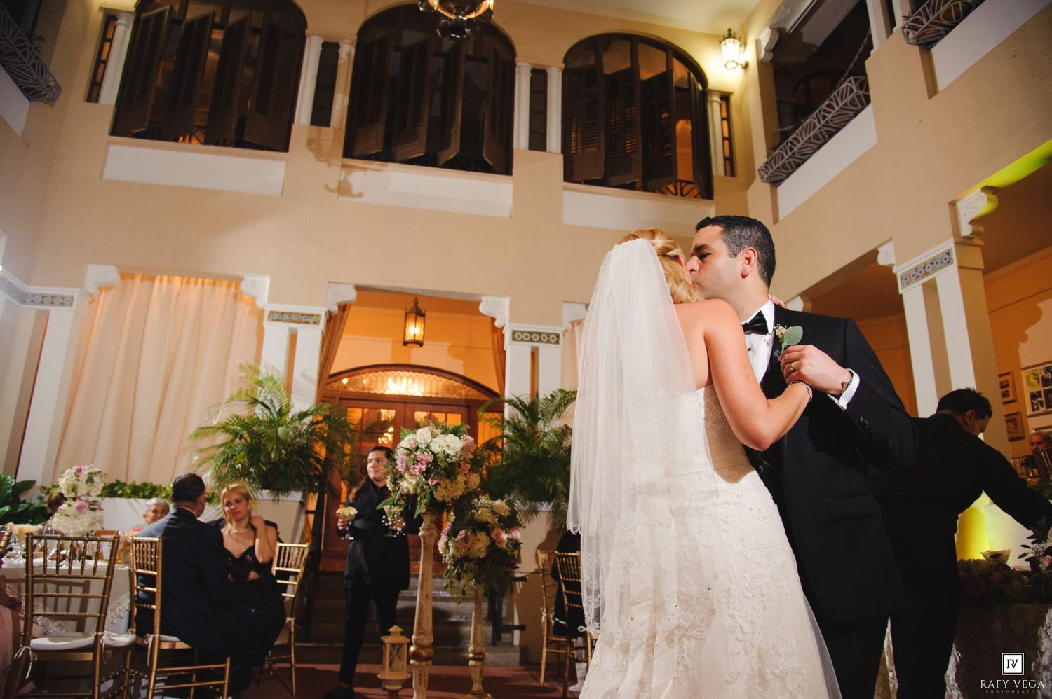 Bride and groom share first dance in elegant ballroom with gold chairs and lush greenery during wedding reception.