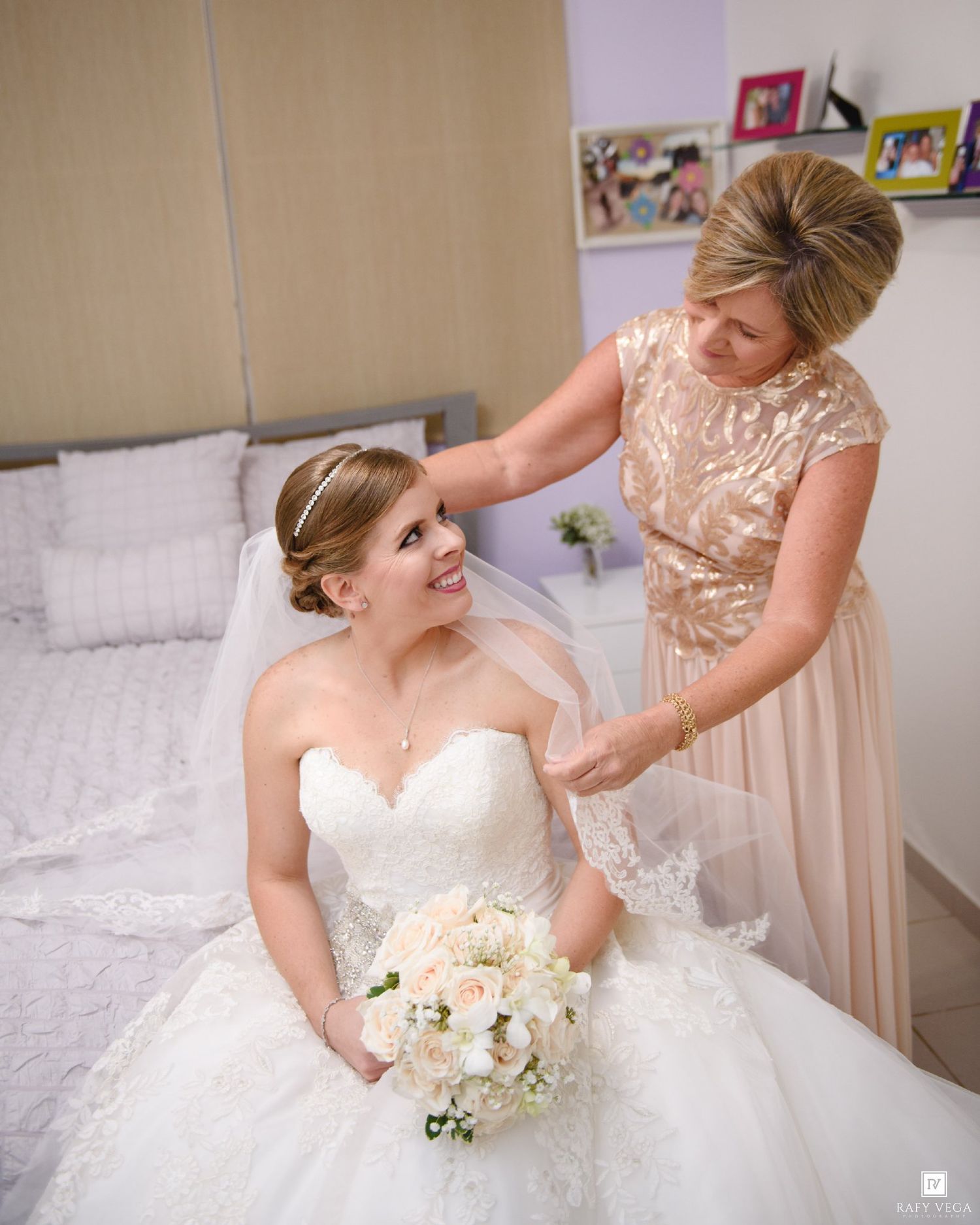 Bride in white ballgown holds bouquet while woman in gold dress adjusts her veil in a bright bedroom.