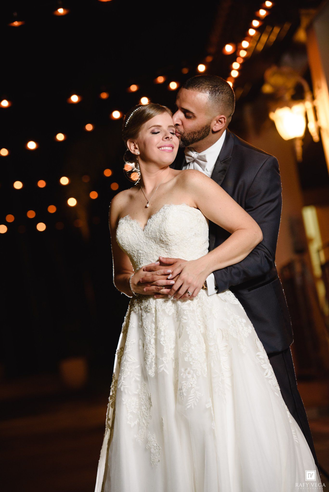 Romantic bride and groom embracing at night under warm string lights, groom kissing bride's cheek in wedding attire.