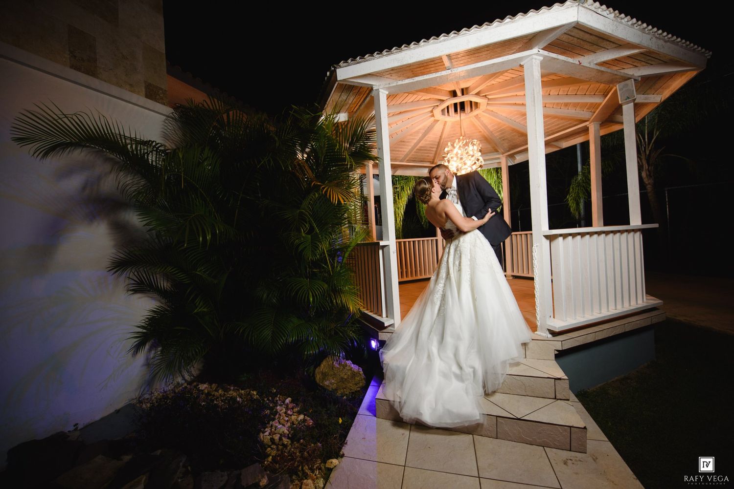 Bride and groom share romantic moment under lit gazebo by pool at night.
