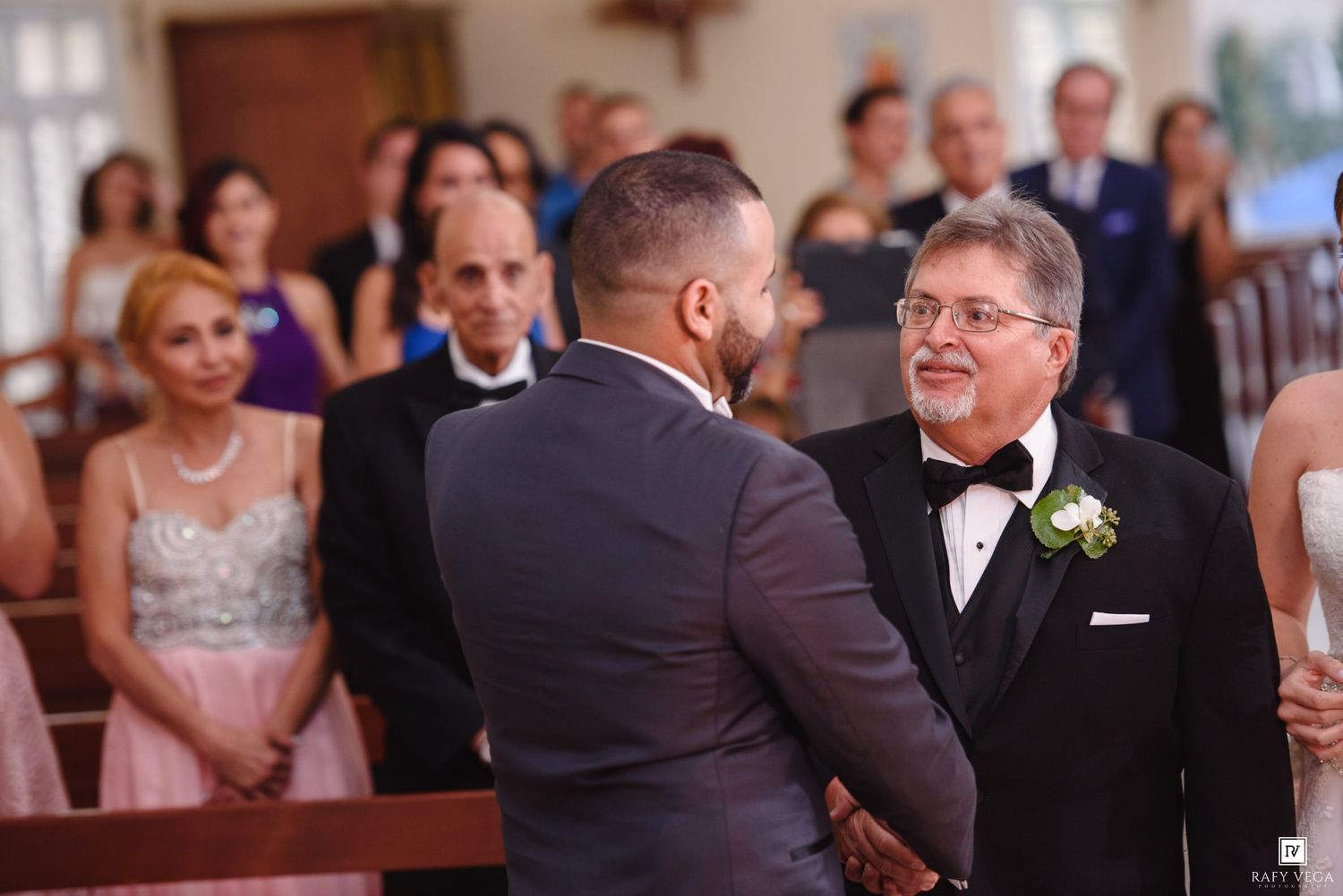 Two men in formal attire shake hands at a wedding ceremony with guests seated in the background.