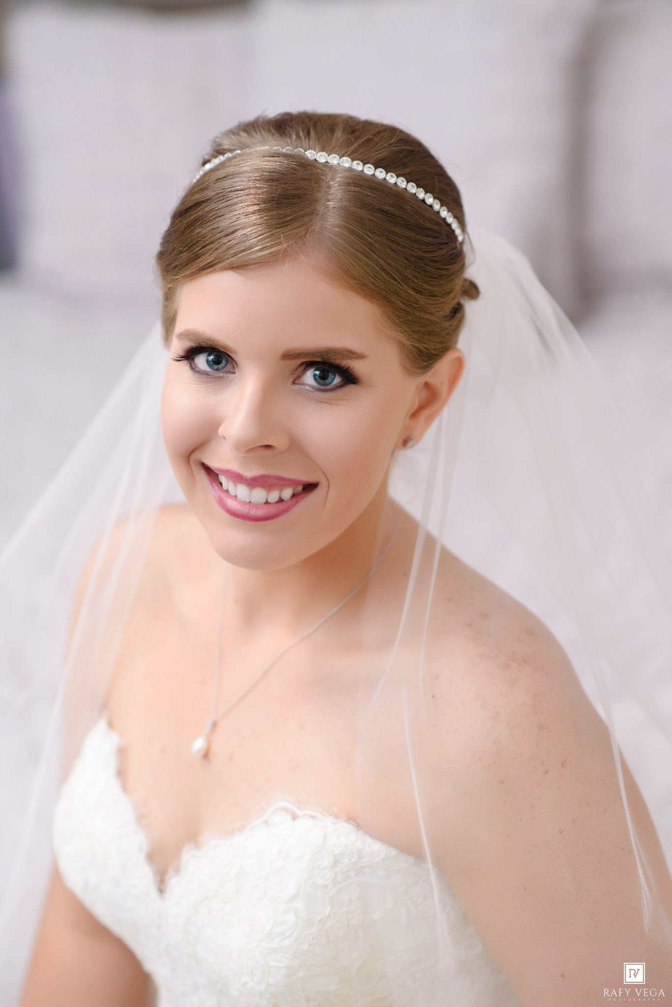Smiling bride wearing a white lace gown, pearl headband, and veil on her wedding day.