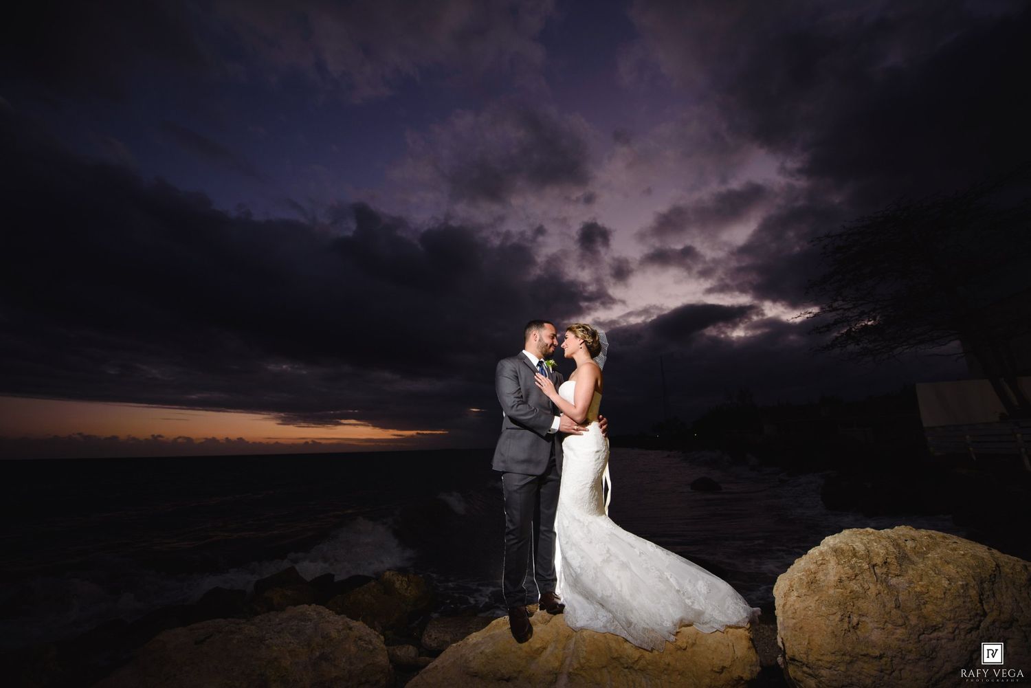 Couple sharing a romantic kiss on rocky shoreline at sunset with dramatic stormy sky backdrop on their wedding day.