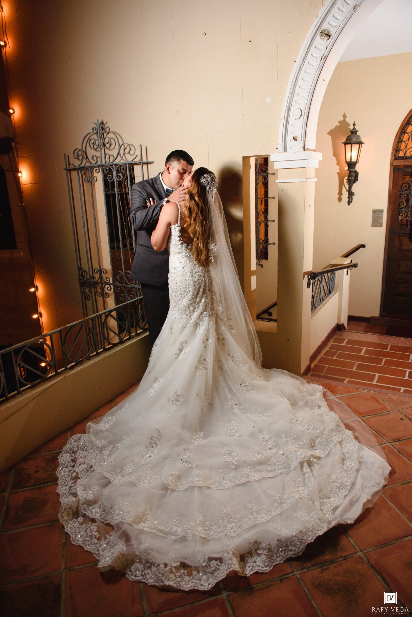 Bride in lace mermaid gown with cathedral veil kissing groom in elegant venue hallway.