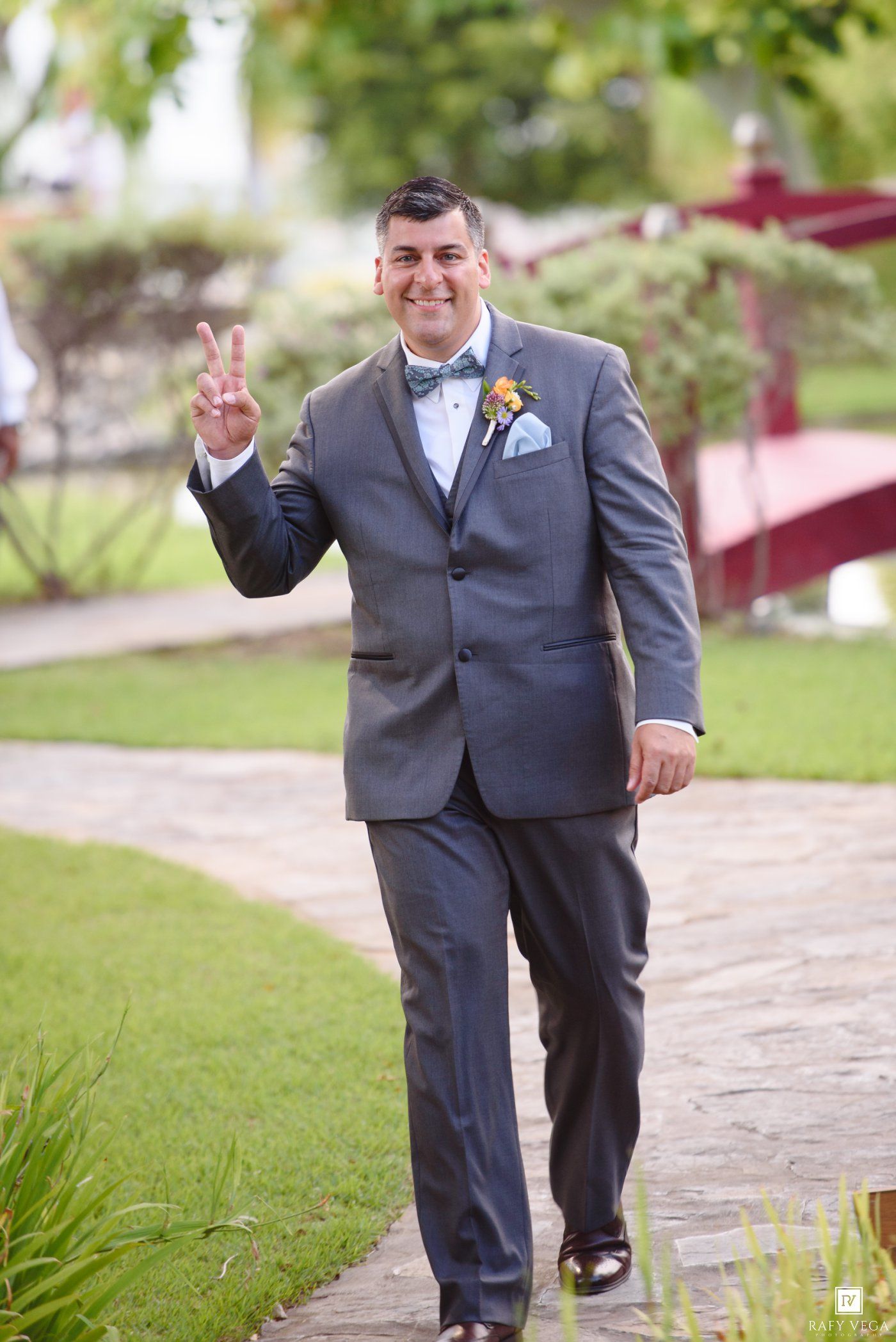 Smiling groom in gray suit with bow tie and boutonniere making peace sign while walking on garden pathway.